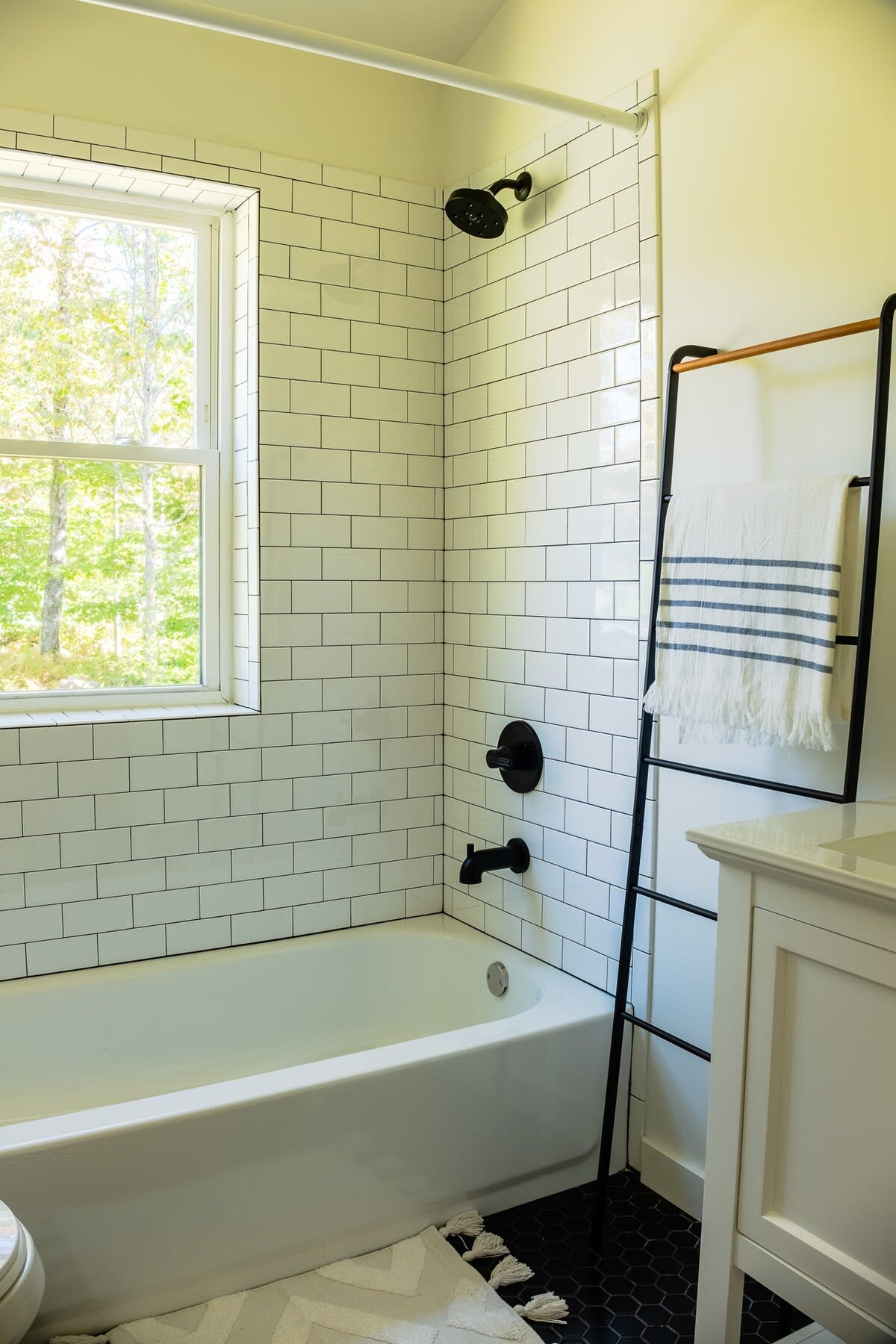 Contemporary bathroom with white subway tiled walls, a white bathtub, and matte black fixtures, including a shower head and faucet. A black ladder-style towel rack holds a striped towel, and a white vanity is partially visible.
