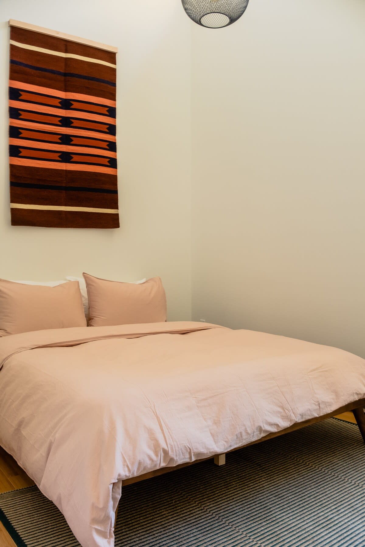 A close-up of a bed with light pink bedding and a striped area rug, with a colorful woven tapestry hanging above the bed on a light-colored wall.