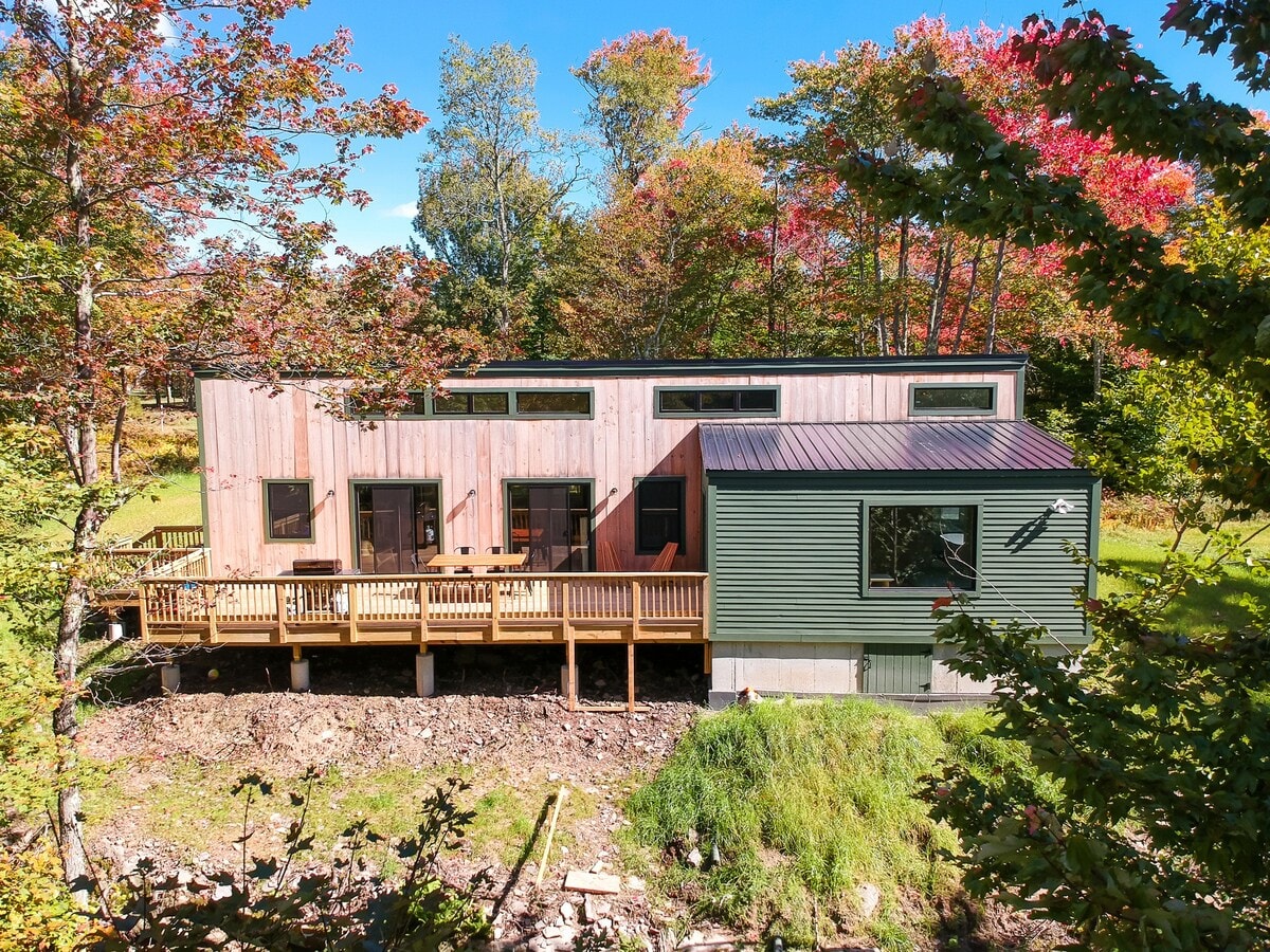 Rear view of the modern cabin showcasing its multi-toned siding, a large wooden deck with steps leading down, and a backdrop of vibrant autumn trees. The house features multiple windows and a sloped roofline.