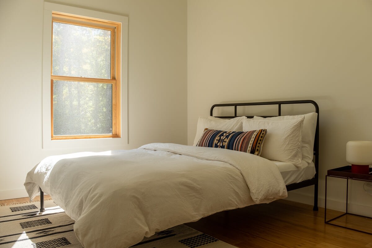Bright bedroom with a black metal bed frame, white bedding, and a colorful patterned throw pillow, set against light-colored walls and hardwood floors. A large window offers a view of trees, filling the room with natural light.