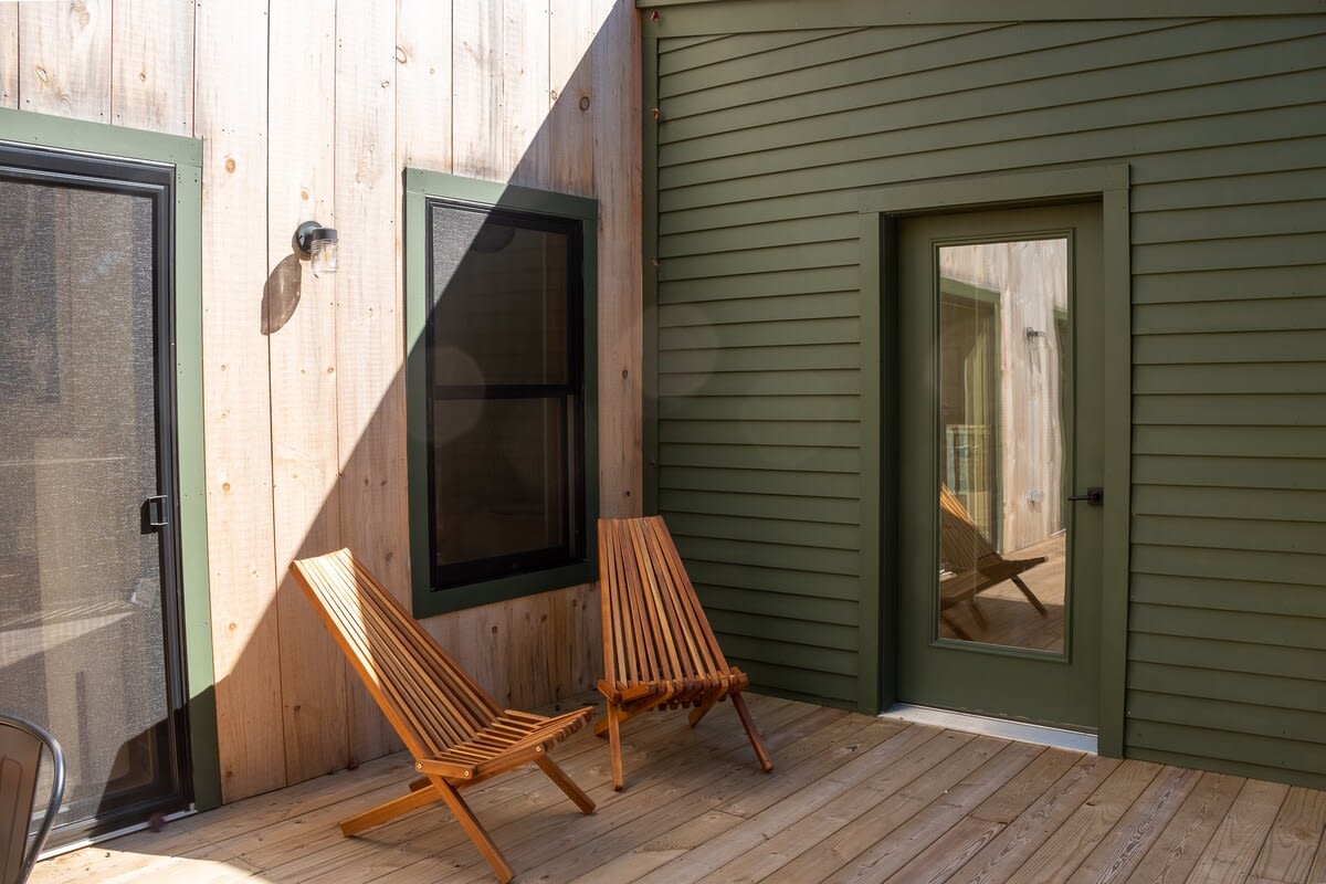 Section of a wooden deck with two slatted wooden lounge chairs, positioned against the house exterior which features both natural wood and dark green siding. A glass door and window are also visible.
