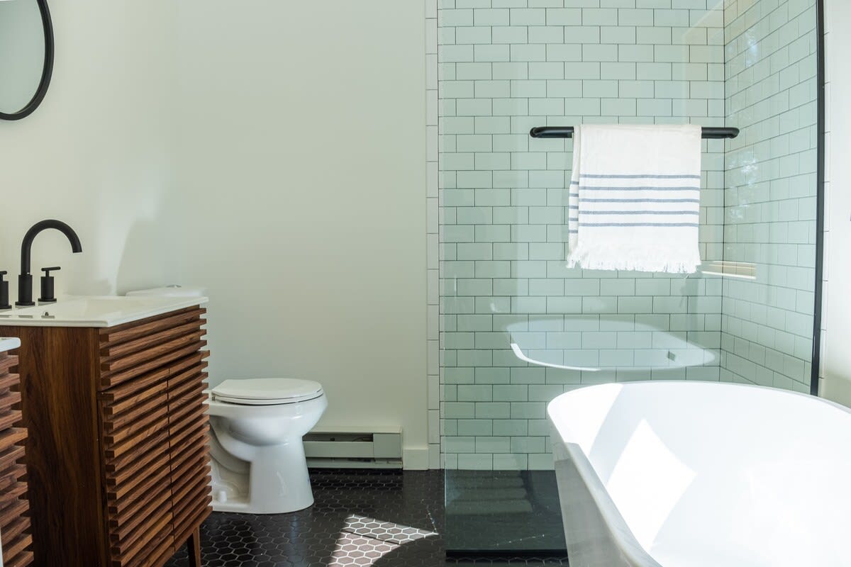 A modern bathroom featuring a dark wood vanity with a black faucet, a white toilet, a freestanding white bathtub, and a walk-in shower with light green subway tiles and a glass enclosure, all on a dark hexagonal tiled floor.