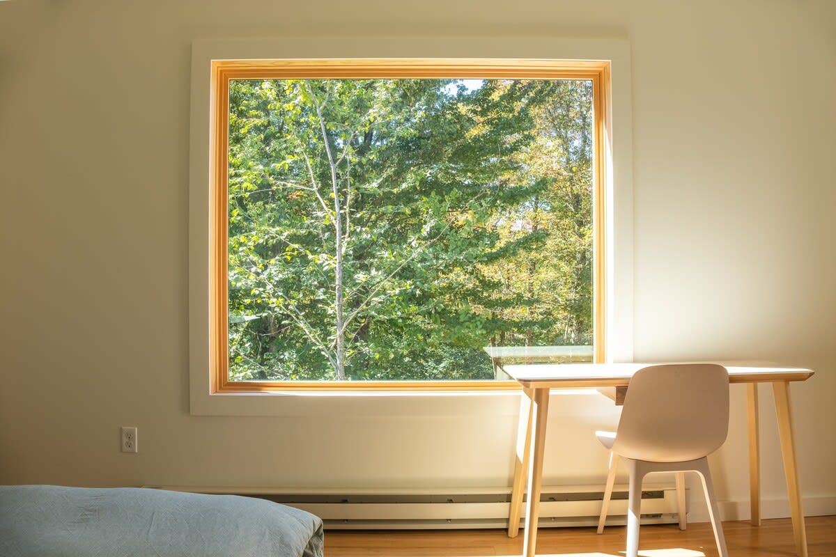 A close-up of a large window in a bedroom, framed in light wood, offering a vibrant view of green trees, with a simple light wood desk and chair positioned beneath it.