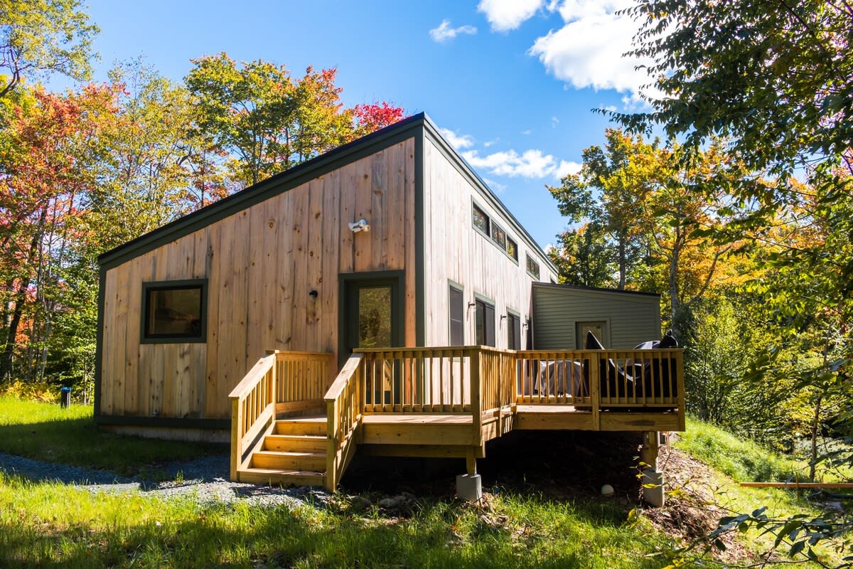 Modern cabin-style house with natural wood siding and a dark green trim, featuring a wooden deck with stairs leading down to a grassy yard. The house is nestled among vibrant autumn trees under a clear blue sky.