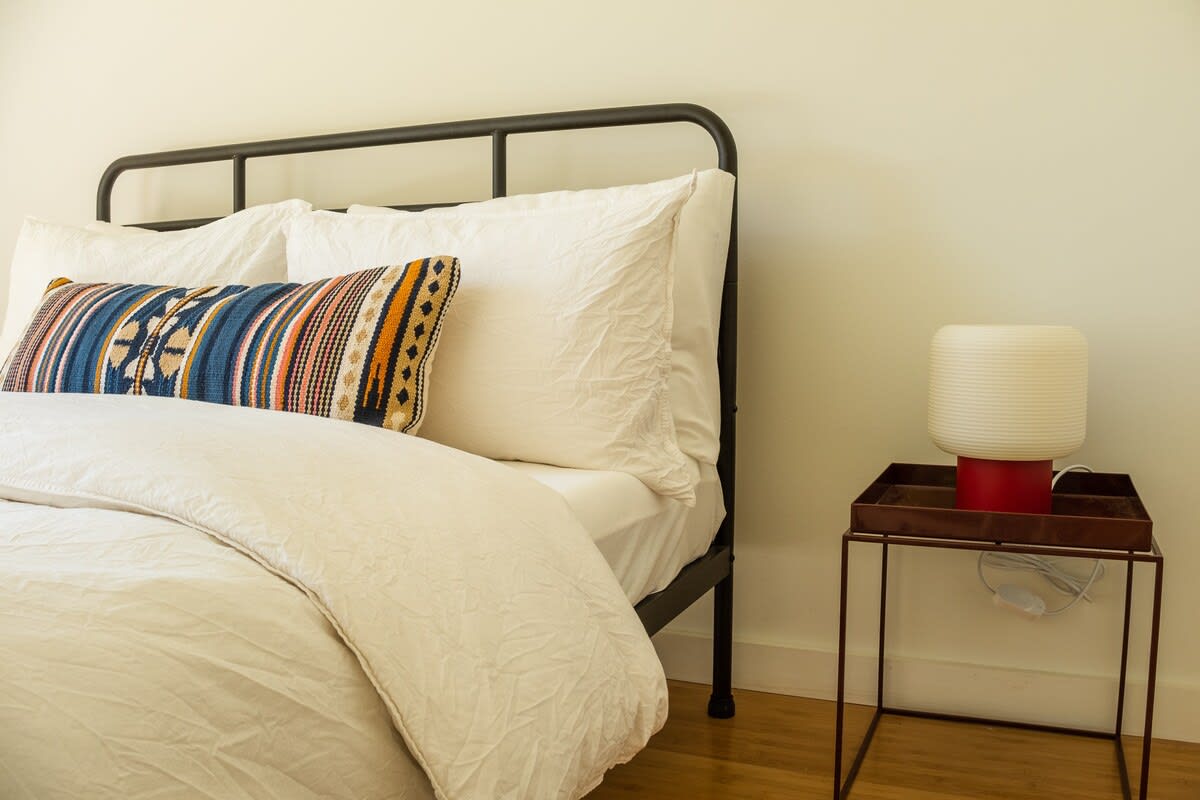 Close-up of a cozy bedroom featuring a black metal bed frame with white bedding and a vibrant, striped throw pillow. A modern nightstand with a red base and a ribbed white lamp sits beside the bed.