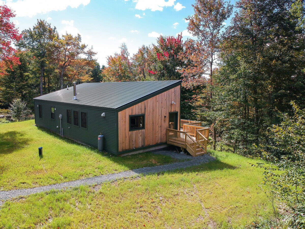 Aerial view of a contemporary house with a dark green metal roof and a mix of natural wood and dark green siding, situated on a grassy hill surrounded by dense, colorful autumn foliage. A gravel path leads to the house.