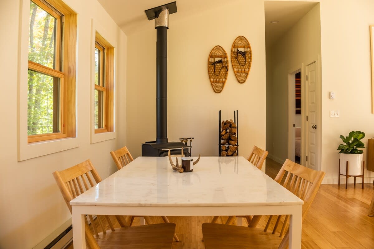 A rustic dining area with a white marble-top dining table, wooden chairs, a black wood-burning stove with a tall pipe, and two decorative snowshoes mounted on the wall, with a view of large windows.