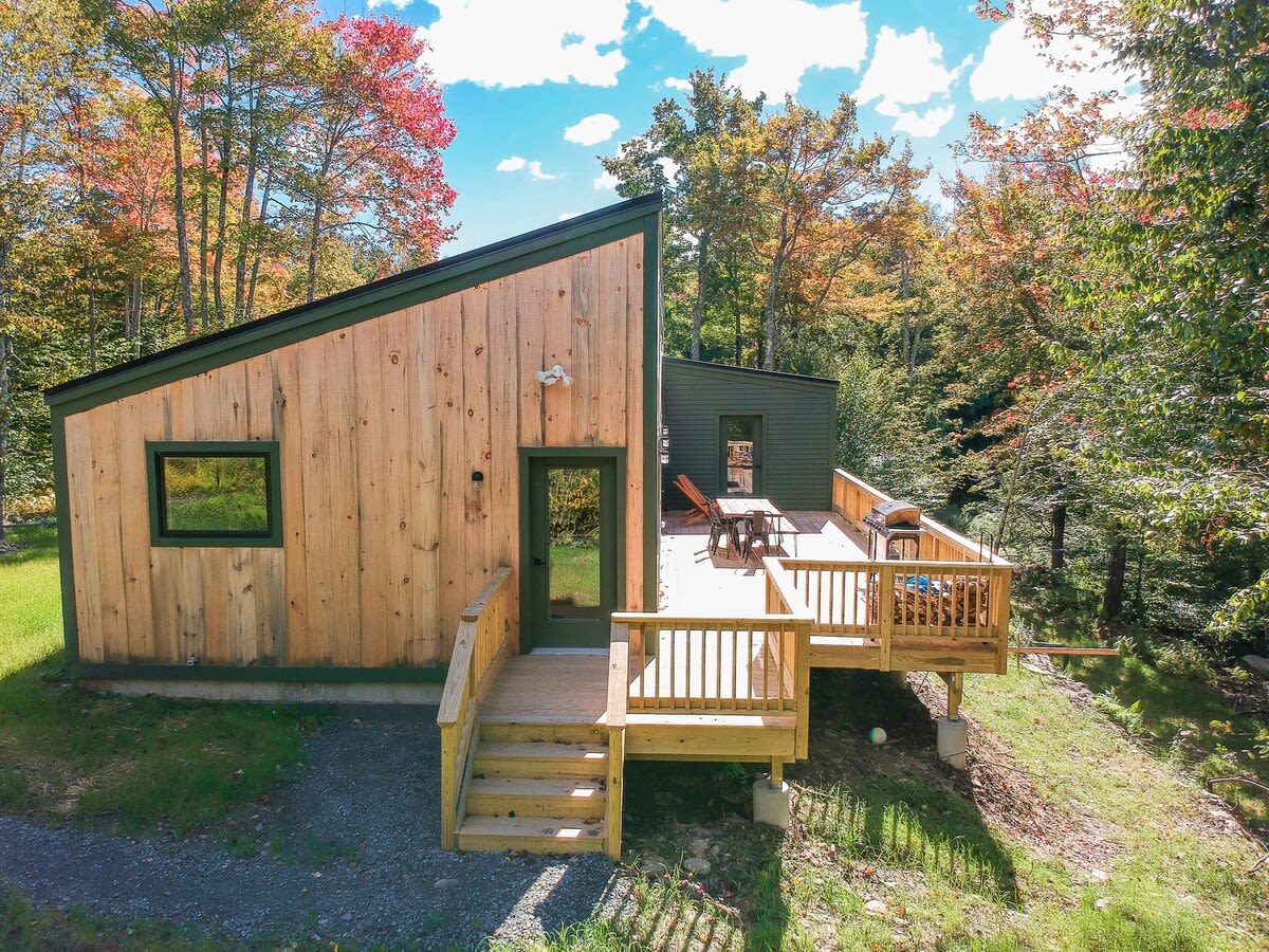 Rear exterior of the modern cabin featuring natural wood siding and dark green accents, with a spacious wooden deck extending from the house. The deck has outdoor seating and is surrounded by colorful autumn trees.