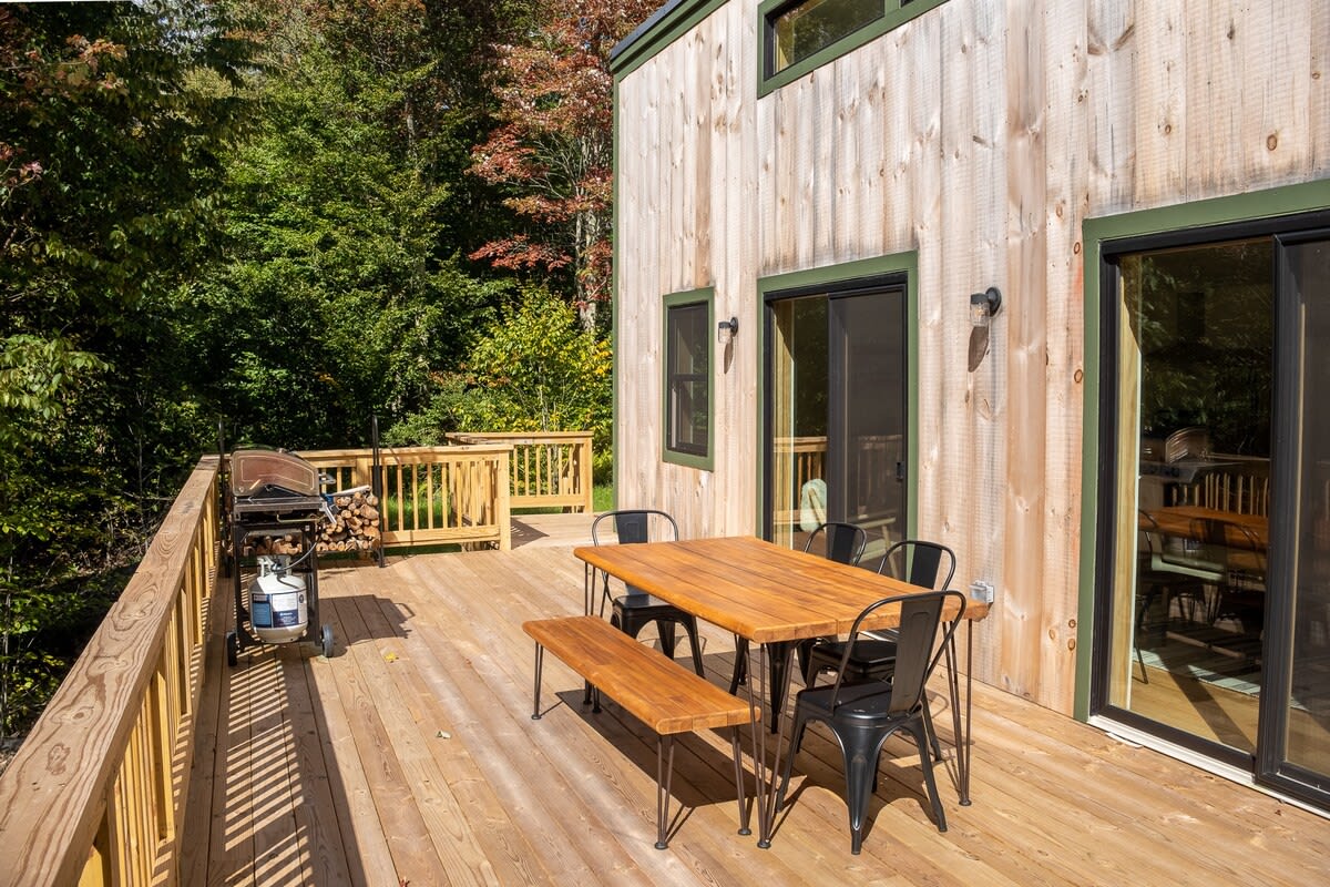 Spacious wooden deck featuring an outdoor dining table with a wooden top and black chairs, a matching bench, and a BBQ grill. The deck is surrounded by a wooden railing and overlooks a wooded area.