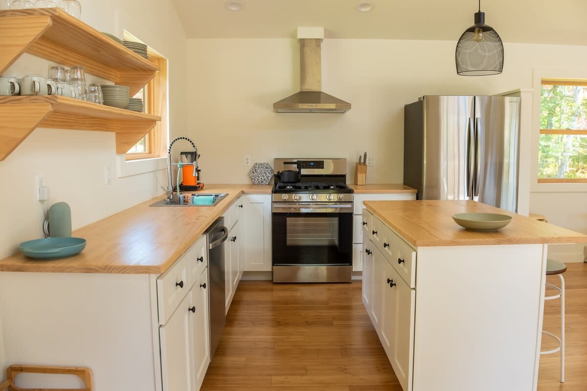 A modern kitchen with light wood countertops, white shaker cabinets, stainless steel appliances including a gas range and refrigerator, a sink under a window, and open wooden shelving.