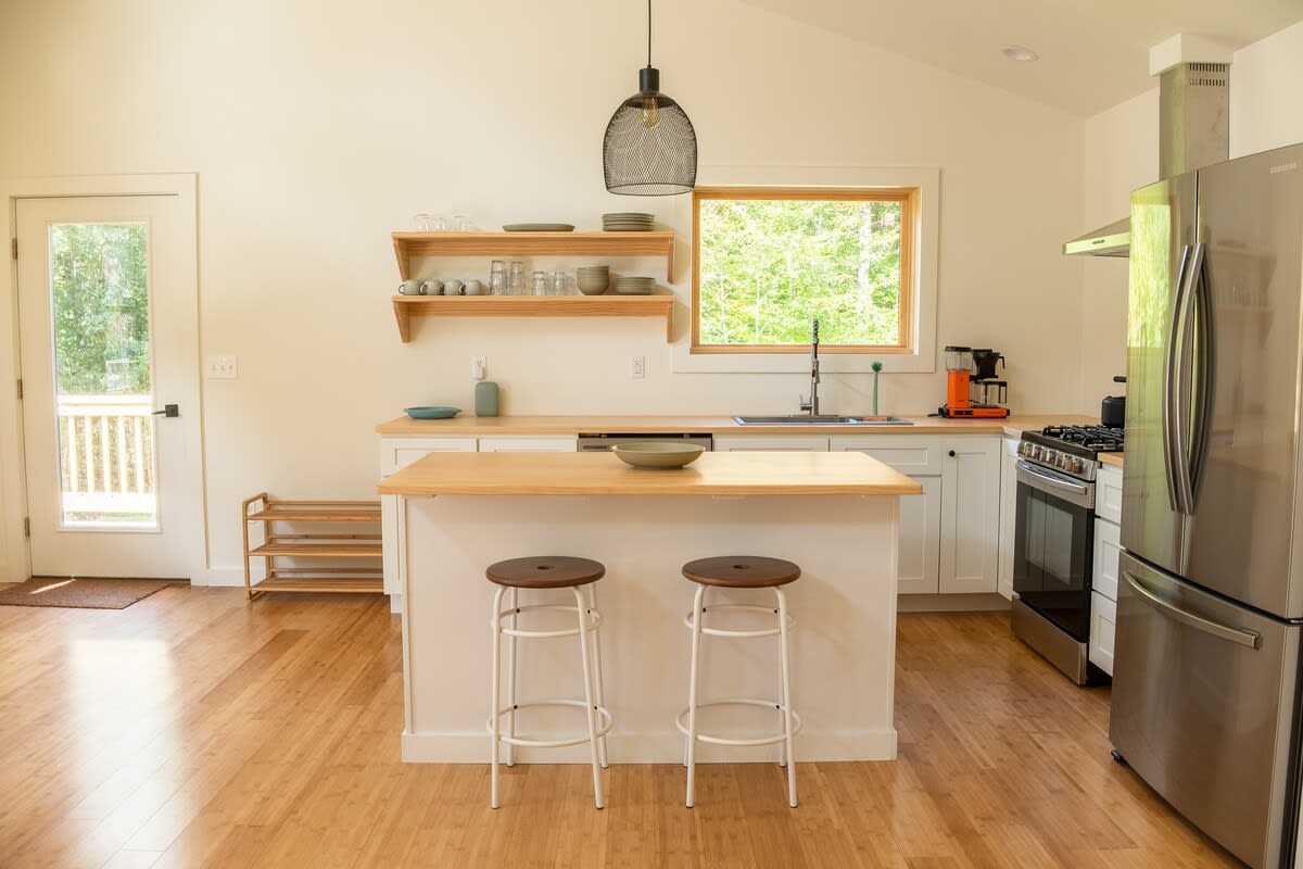 A bright kitchen featuring a white island with two dark-seated bar stools, light wood countertops, white cabinets, stainless steel appliances, and open wooden shelves, with a view towards a glass-paned door.