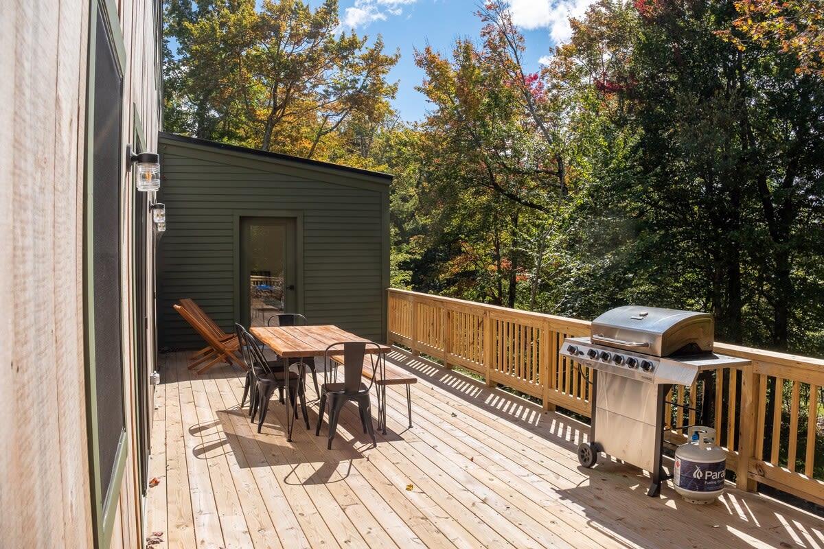 Expansive wooden deck with a large stainless steel BBQ grill and a propane tank, alongside an outdoor dining set. The house exterior features both natural wood and dark green siding, surrounded by vibrant autumn trees.