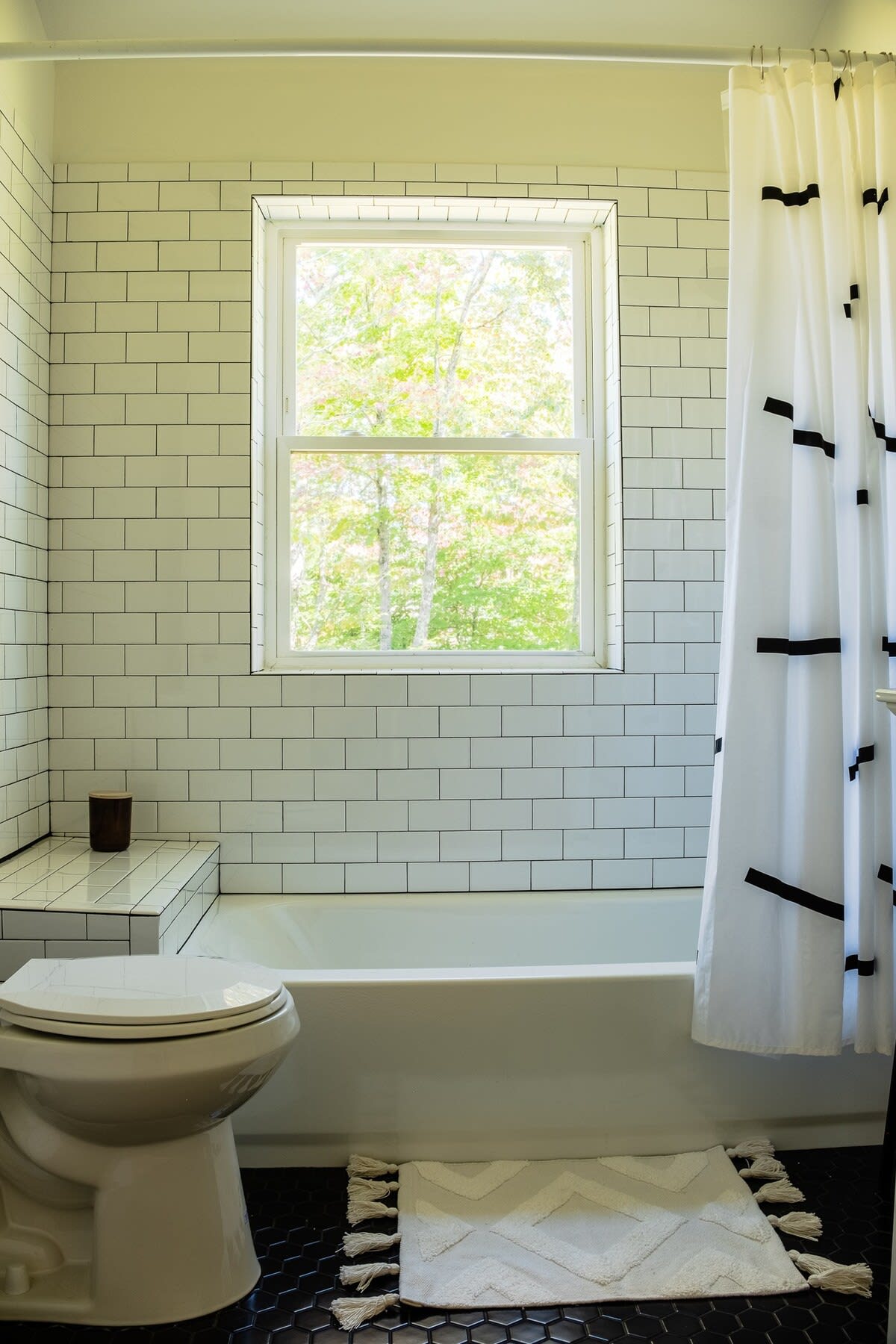Modern bathroom featuring white subway tiles with dark grout, a white bathtub, and a toilet, illuminated by natural light from a window overlooking lush greenery. A white bath mat with a geometric pattern is on the dark hexagonal floor tiles.