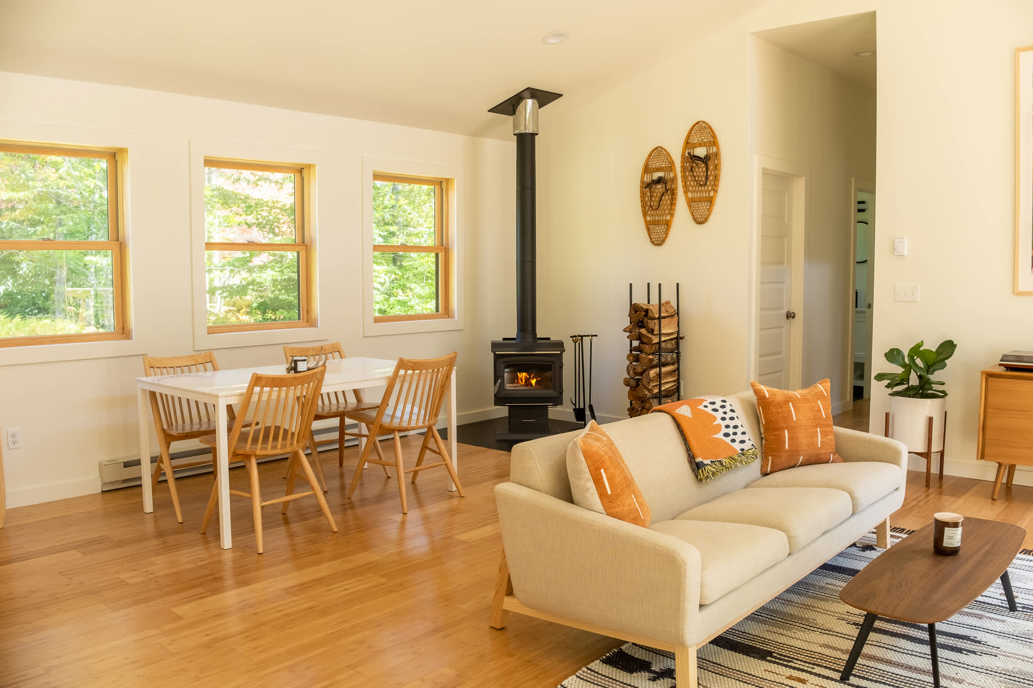 Open-concept living and dining area featuring a modern wood-burning stove, a light-colored sofa with accent pillows, and a white dining table with wooden chairs. Multiple windows offer views of the surrounding greenery.