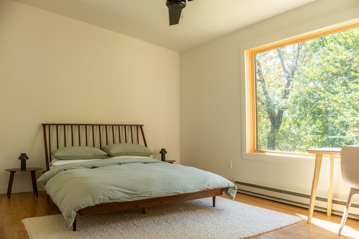 A serene bedroom with a dark wood bed frame, light green bedding, two small round side tables with lamps, a large window overlooking green trees, and a light-colored area rug on hardwood floors.