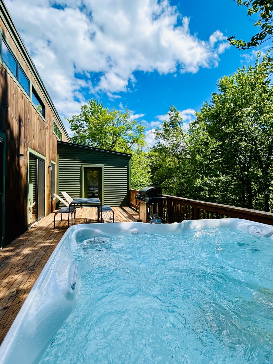 Outdoor hot tub with bubbling water, situated on a wooden deck surrounded by lush green trees under a bright blue sky with white clouds. The side of the house with wooden siding is visible.