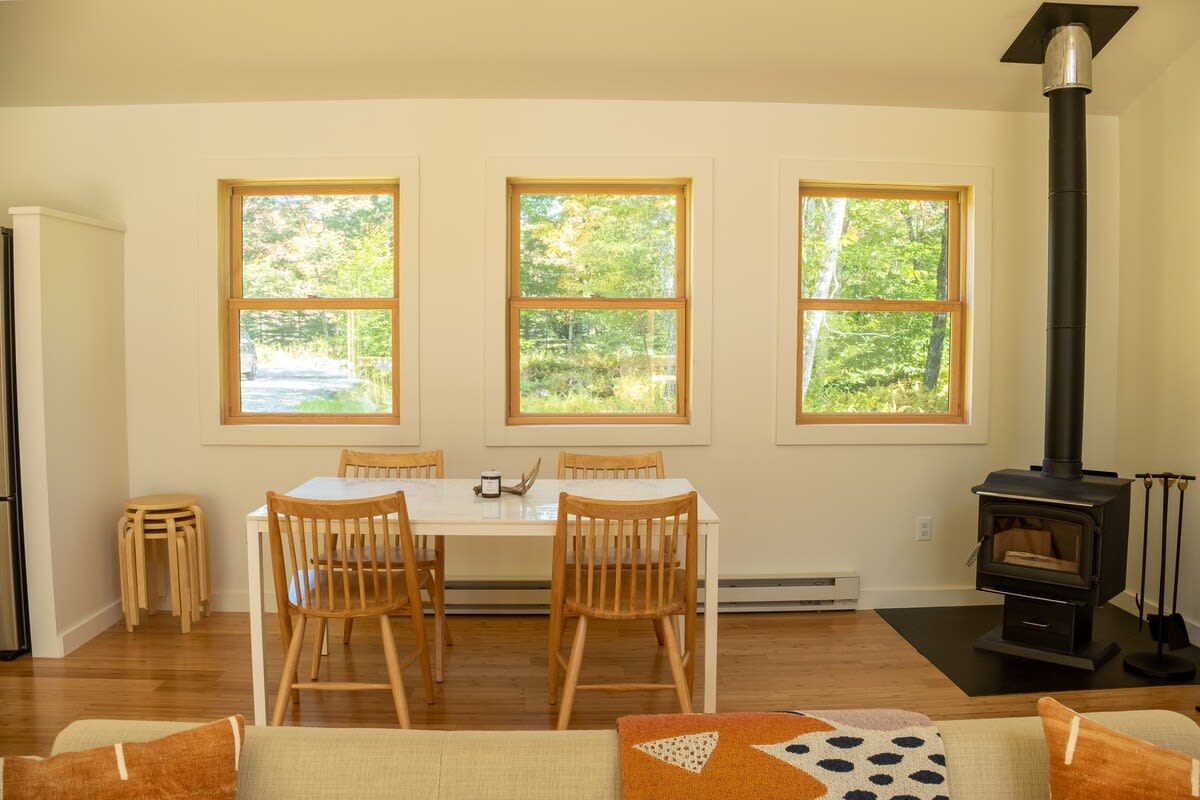 A dining area with a white dining table and wooden chairs, positioned in front of three large windows offering a view of lush green trees, adjacent to a black wood-burning stove with a tall pipe.