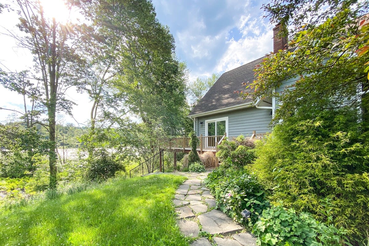 Charming house exterior with light blue siding and a dark roof, nestled among mature trees and lush greenery. A stone pathway leads through a grassy yard towards a deck with a sliding glass door, offering glimpses of a lake in the background.
