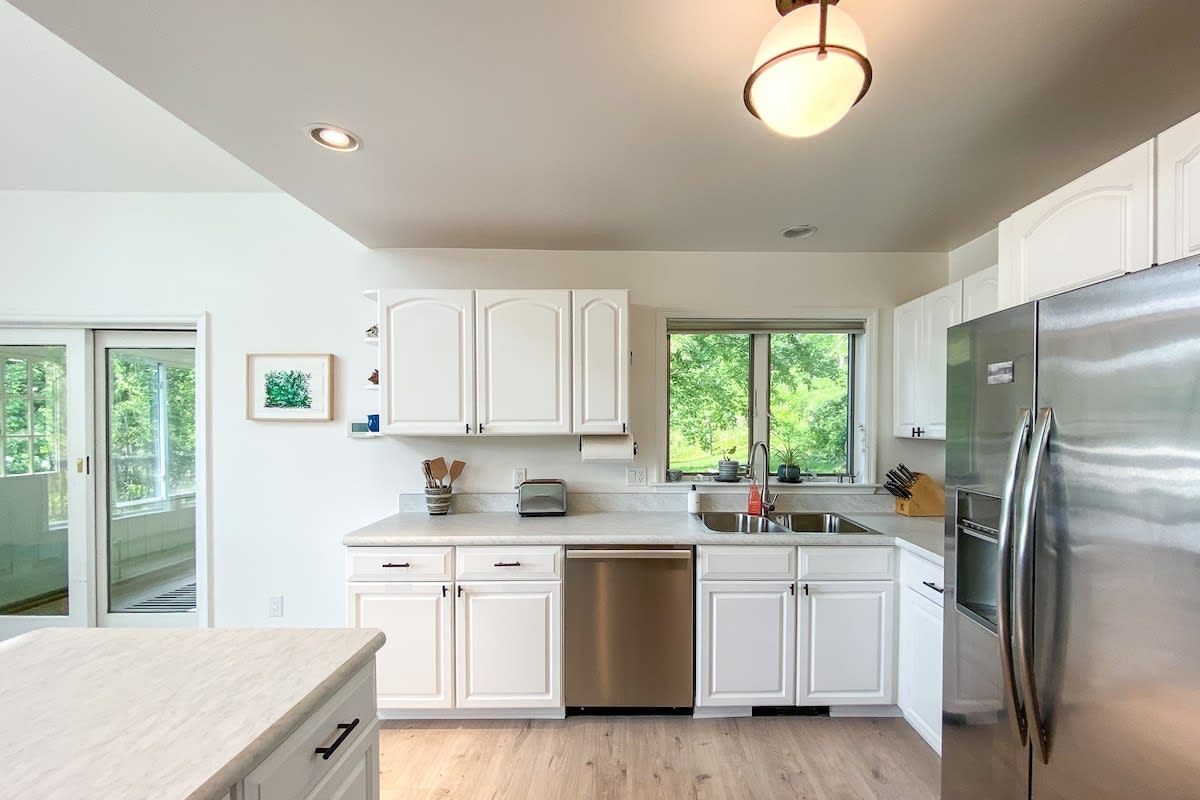 Modern kitchen with white cabinetry, light countertops, and a double sink under a large window offering views of lush greenery. Stainless steel dishwasher and toaster are visible on the counter.