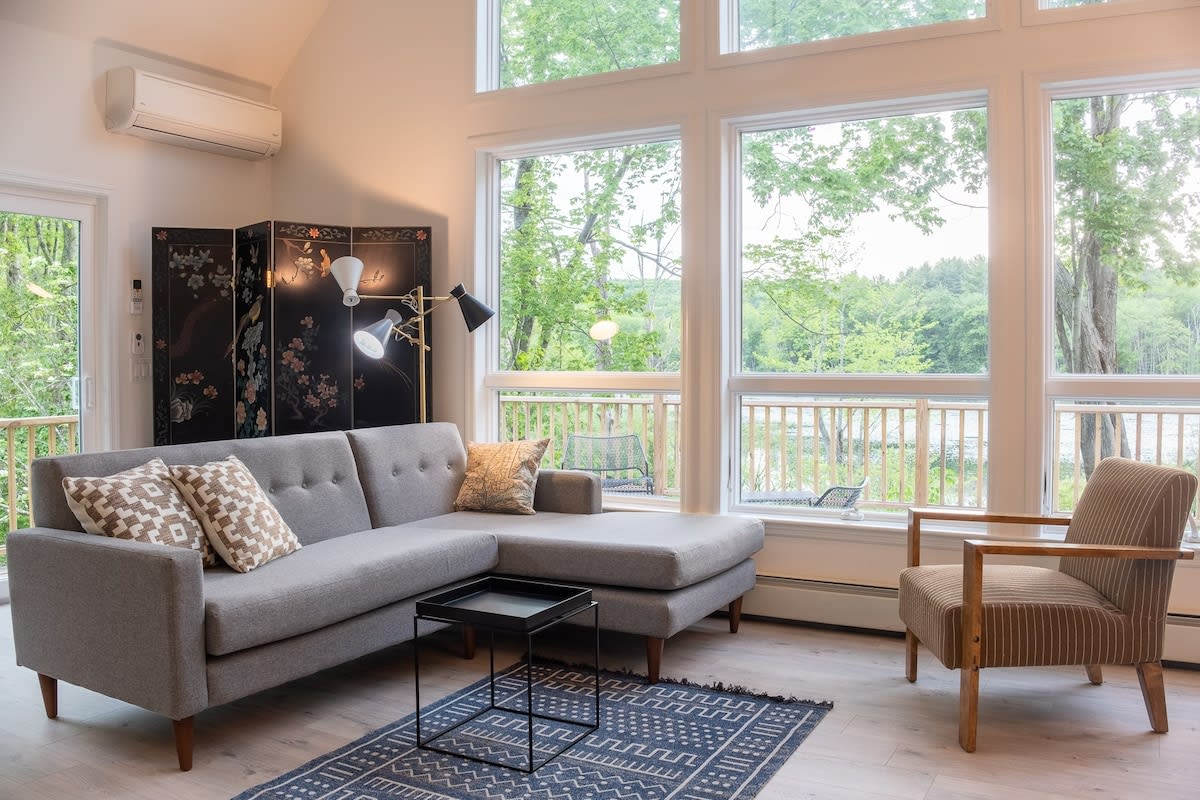 A bright living area showcases a comfortable gray sectional sofa with patterned pillows, a black side table, and a dark blue geometric rug. Large windows offer a view of the outdoor deck and surrounding trees, with an air conditioning unit mounted above.