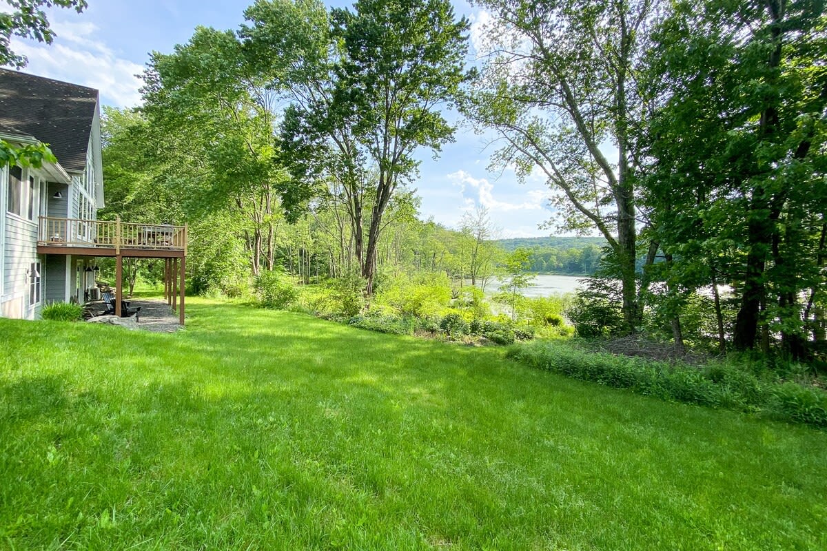 Spacious, well-maintained grassy yard surrounded by dense green trees, with a clear view of a serene lake in the distance. The side of the house with a raised deck is visible on the left.