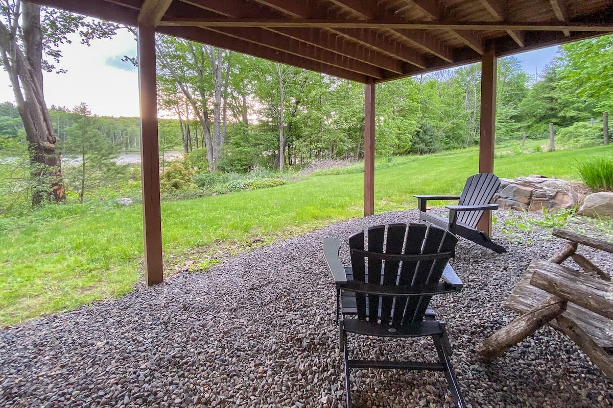 A shaded outdoor patio area beneath the main deck features dark Adirondack chairs on a gravel surface. It offers a serene view of the grassy backyard and distant trees, providing a quiet spot for relaxation.