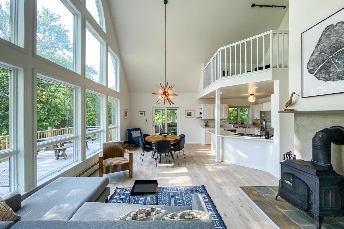 An expansive open-concept living and dining area boasts a vaulted ceiling, a gray sectional sofa, and a round wooden dining table. A wood-burning stove sits beside a stone fireplace, with a modern kitchen visible in the background and large windows overlooking the deck and lake.