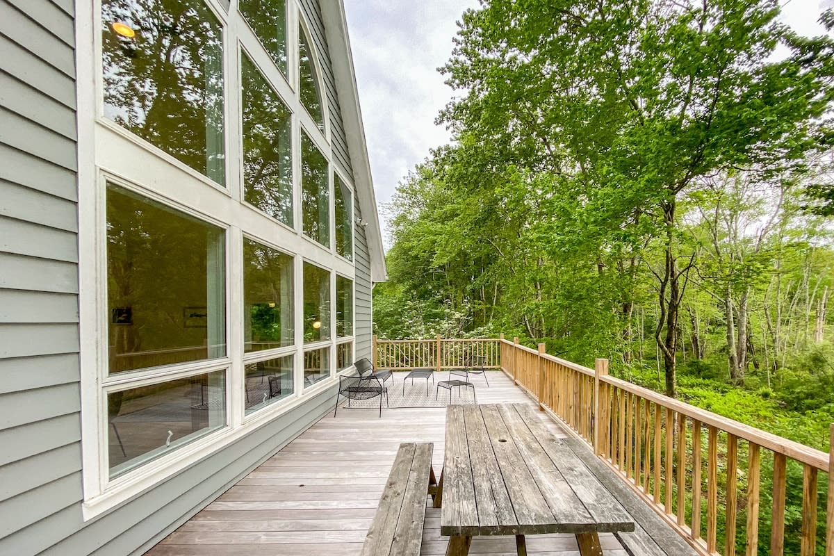 Expansive wooden deck with a rustic picnic table and metal chairs, offering panoramic views of lush green trees and a distant body of water. The house features large, multi-pane windows reflecting the sky.