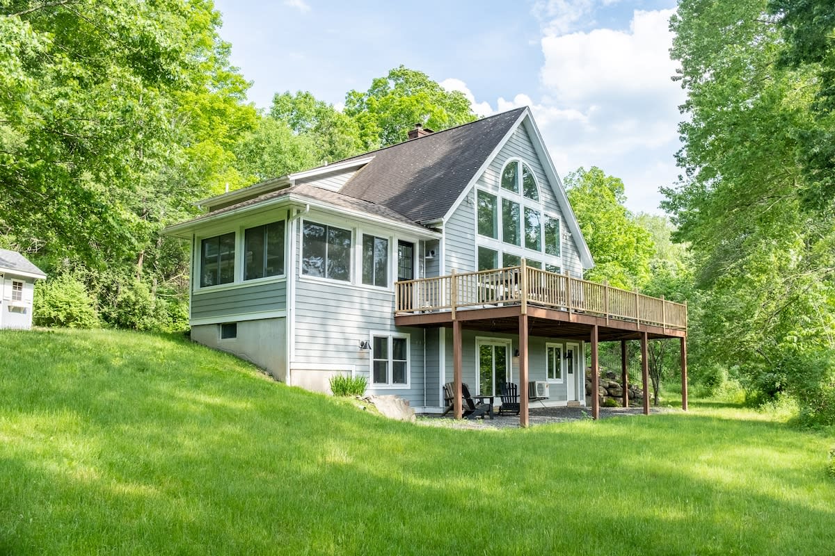 The back of the house features light gray siding, a large wooden deck, and expansive windows that reveal the interior living space. The property is set on a grassy slope surrounded by lush green trees under a partly cloudy sky.