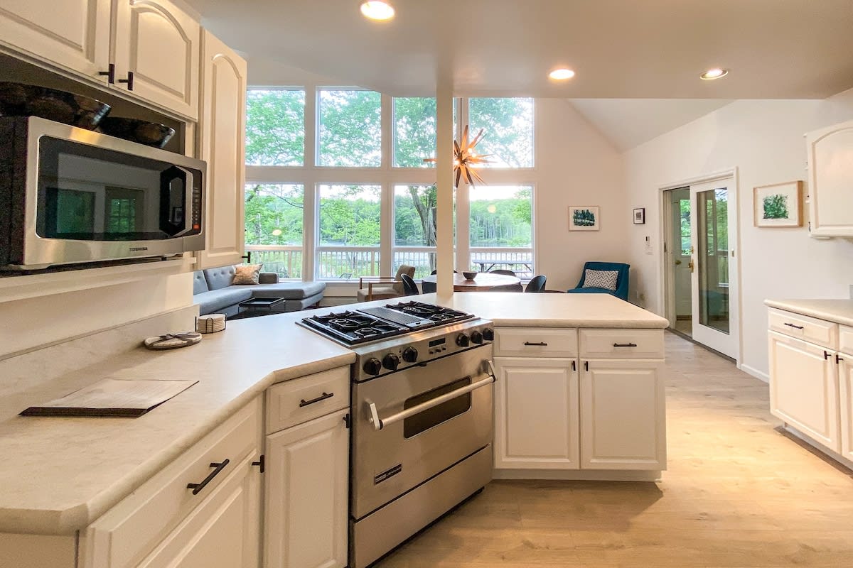Open-concept kitchen with a stainless steel range and a large island featuring light countertops and white cabinets. The background reveals a living area with expansive windows overlooking a lake.
