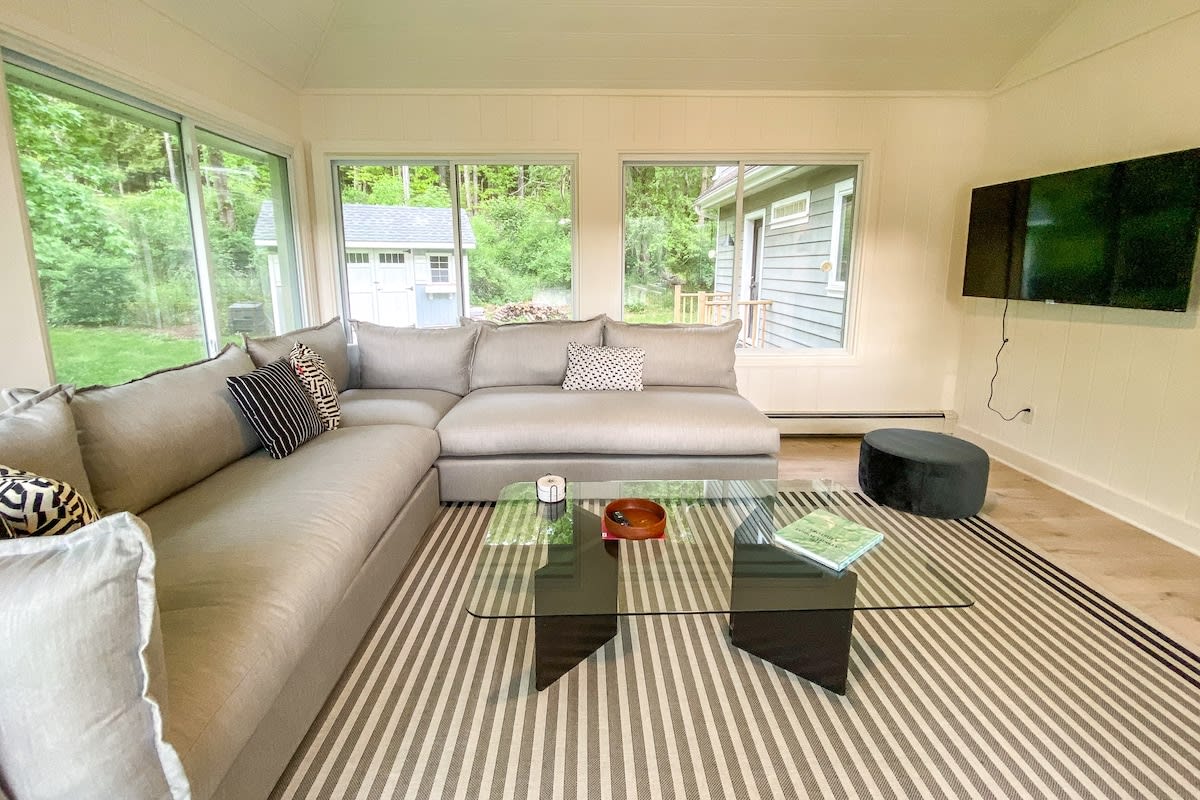 A cozy living space features a large light gray sectional sofa with decorative pillows, a glass coffee table, and a striped rug. Large windows provide views of the surrounding greenery and a nearby building.