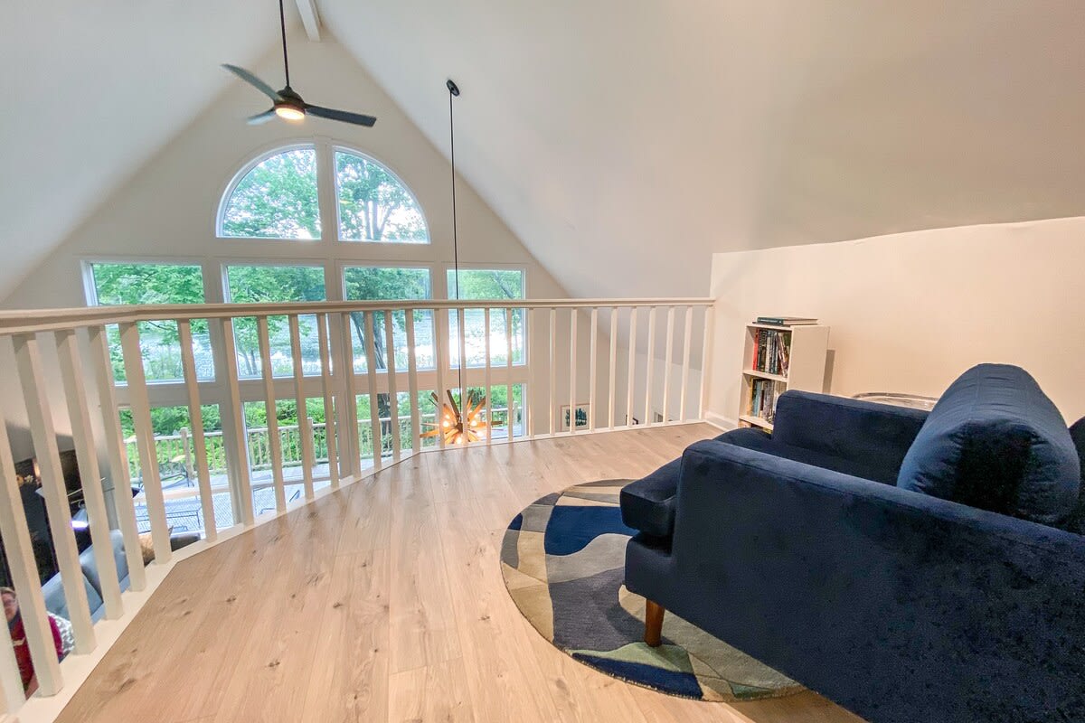 Bright loft area overlooking the main living space, featuring a blue sofa and a round rug. Large windows provide expansive views of the lake and surrounding nature.
