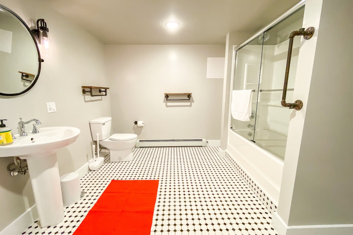 Modern bathroom featuring a white pedestal sink, toilet, and a glass-enclosed shower with industrial-style pipe fixtures. The floor is covered in black and white hexagonal tiles, contrasted by a bright red bath mat.