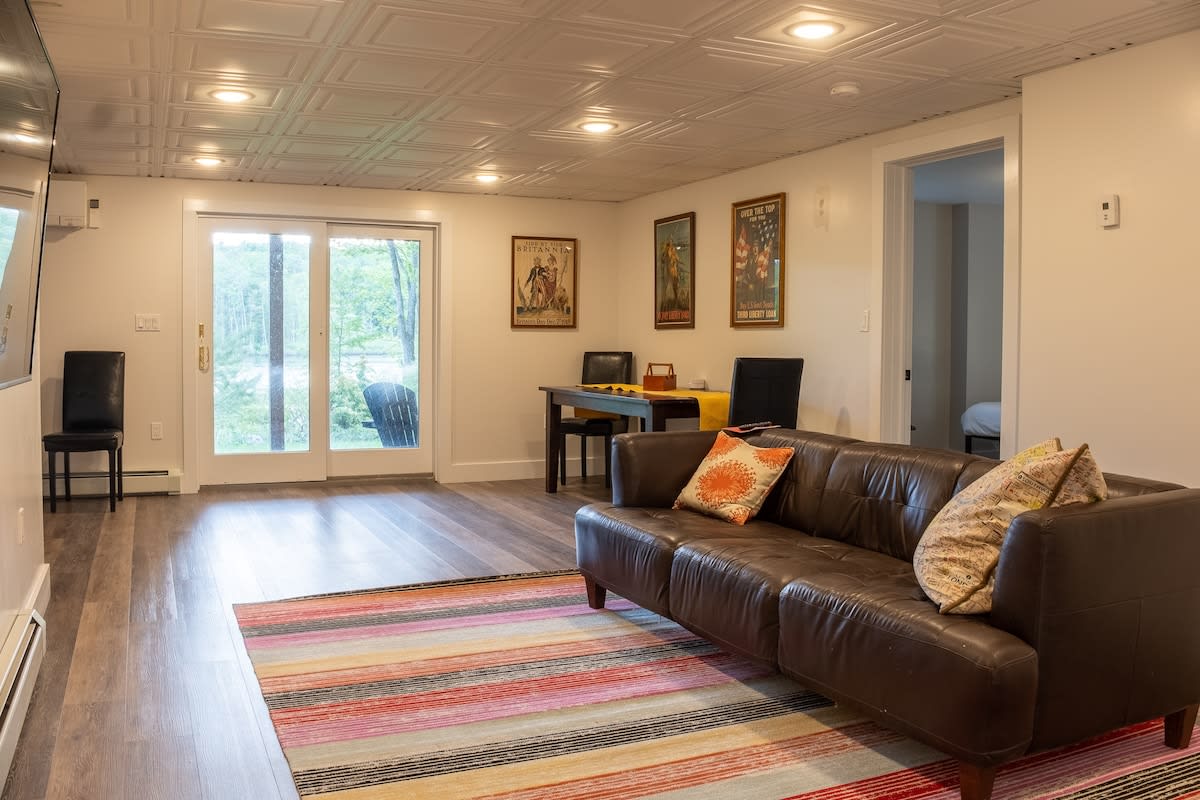 A casual basement living area features a brown leather sofa with colorful pillows, a striped rug, and a small dining or game table. Sliding glass doors open to the outdoors, and framed artwork adorns the walls.