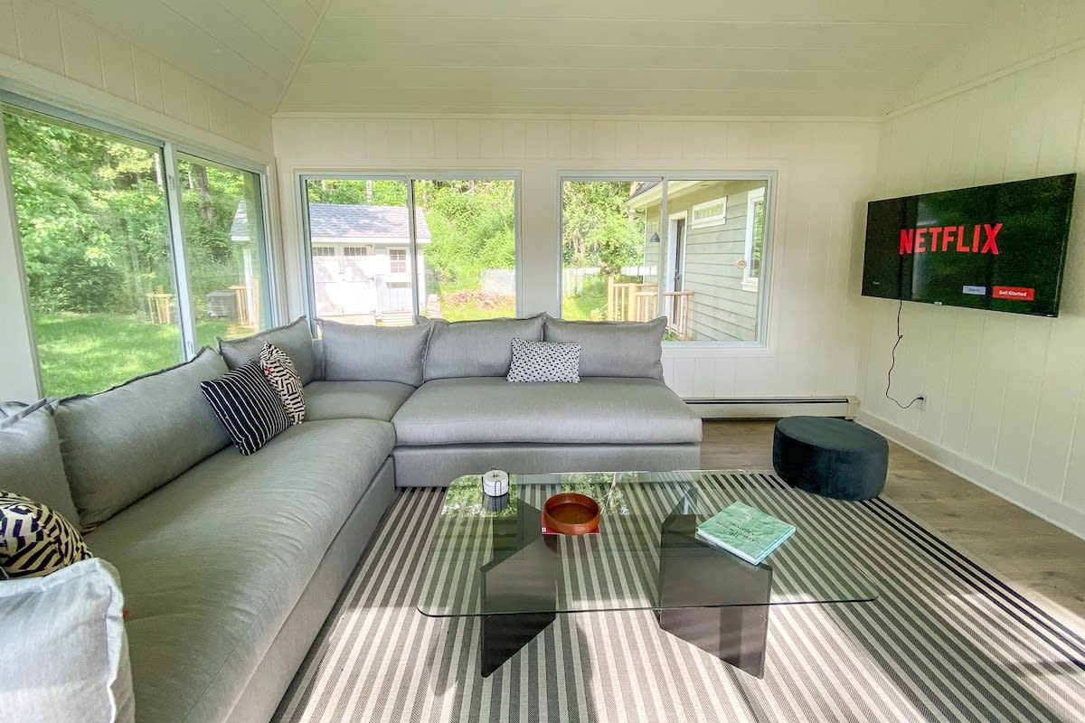 Cozy sunroom living area with a large grey sectional sofa, a glass coffee table on a striped rug, and a wall-mounted TV displaying Netflix. Multiple windows offer views of the surrounding greenery.