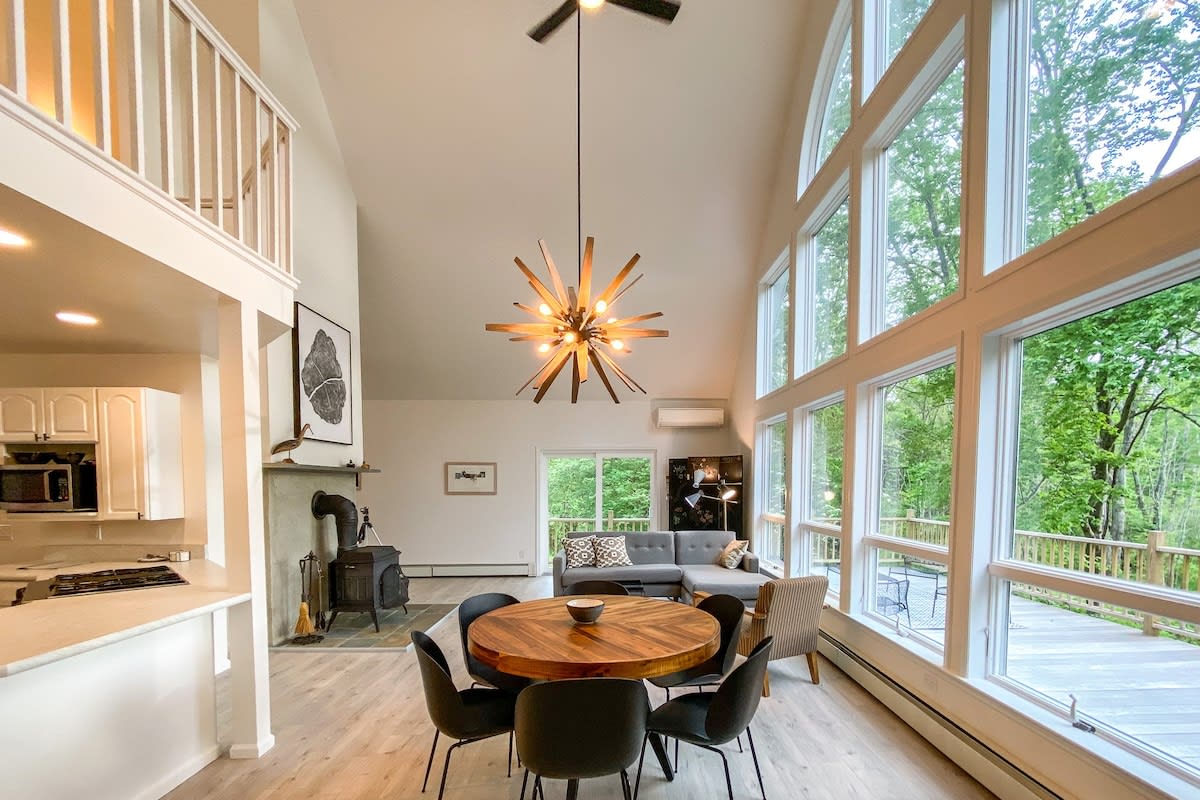 The bright, open-plan living and dining area showcases a striking sputnik chandelier, a round wooden dining table with black chairs, and a gray sectional sofa. A wood-burning stove is integrated into a stone fireplace, and a loft area overlooks the space, with large windows framing the outdoor scenery.