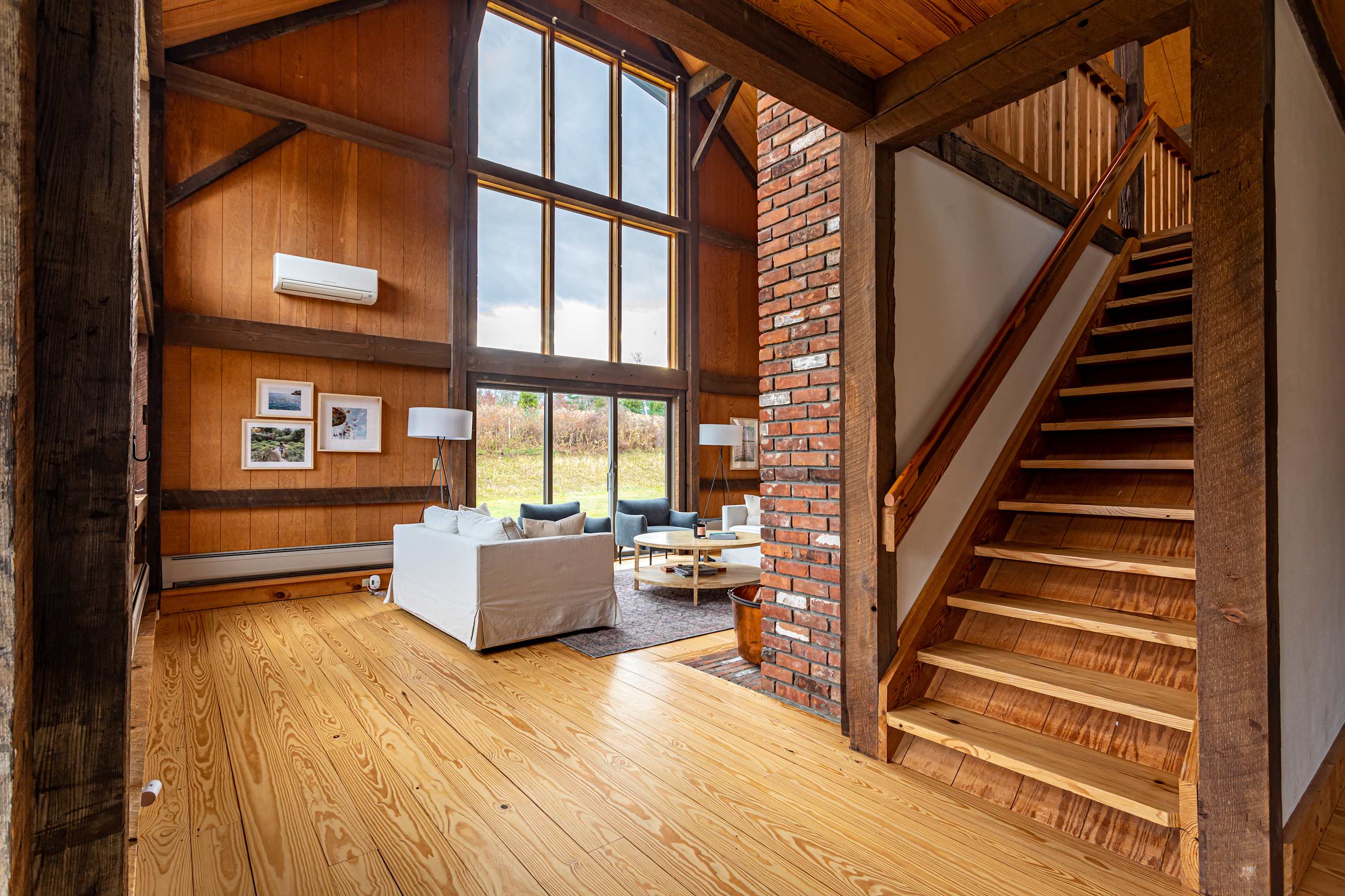 Expansive living room with soaring ceilings, exposed wooden beams, and large windows offering outdoor views. A white sofa faces a coffee table, and a brick fireplace stands next to a wooden staircase.