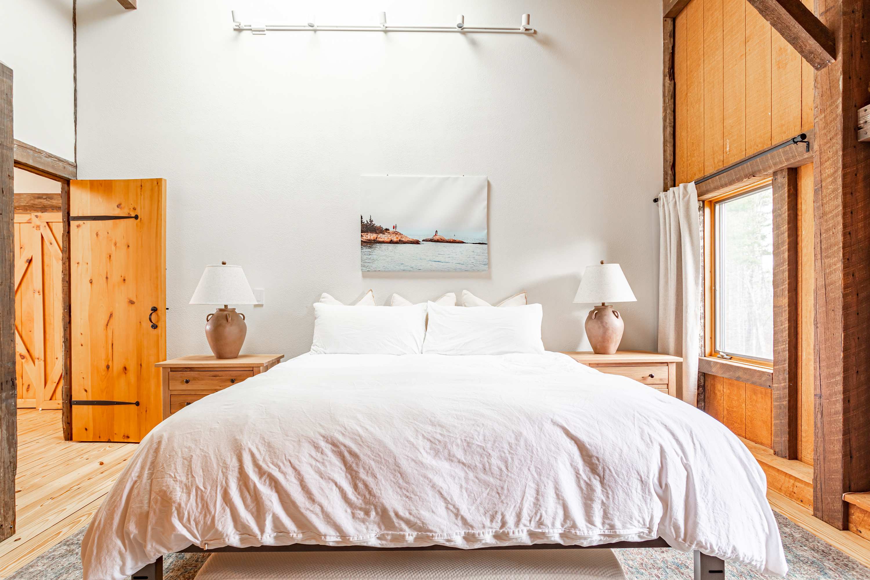 Bright bedroom featuring a king-sized bed with white bedding, flanked by two wooden nightstands with ceramic lamps. A large artwork hangs above the headboard.