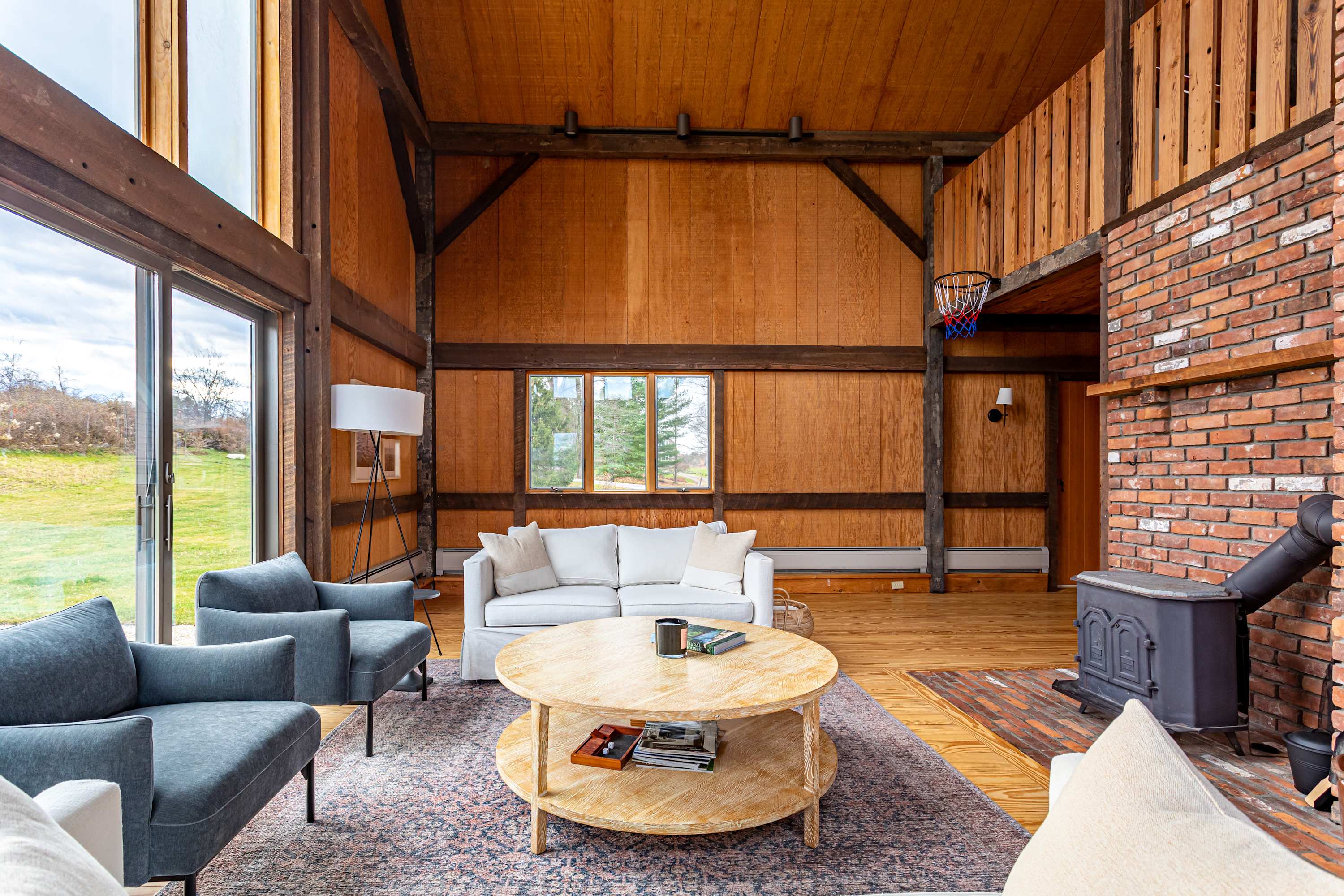 A rustic living room features a white sofa and two gray armchairs arranged around a round wooden coffee table on a patterned rug, with a brick fireplace and wood-burning stove to the right and large glass doors leading to the outdoors on the left. The room has high vaulted ceilings and walls clad in warm-toned wood, with exposed wooden beams. A mini basketball hoop is visible on the upper level.