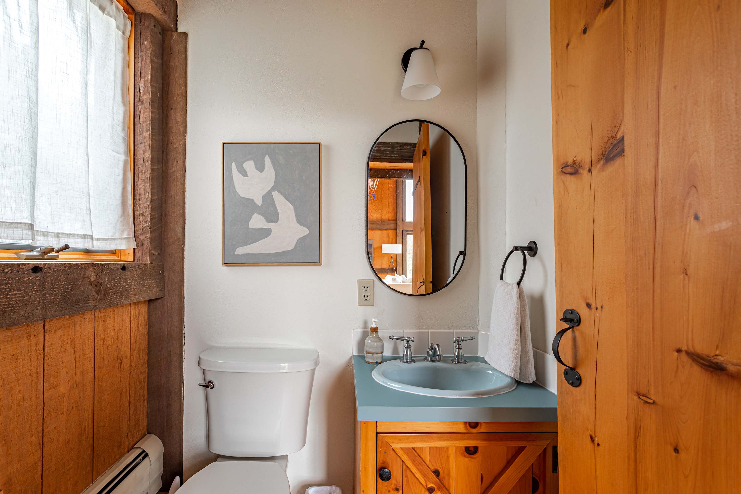 Compact half-bathroom featuring a white toilet, a light wood vanity with a light blue countertop, and an oval black-framed mirror. A modern abstract artwork hangs above the toilet.