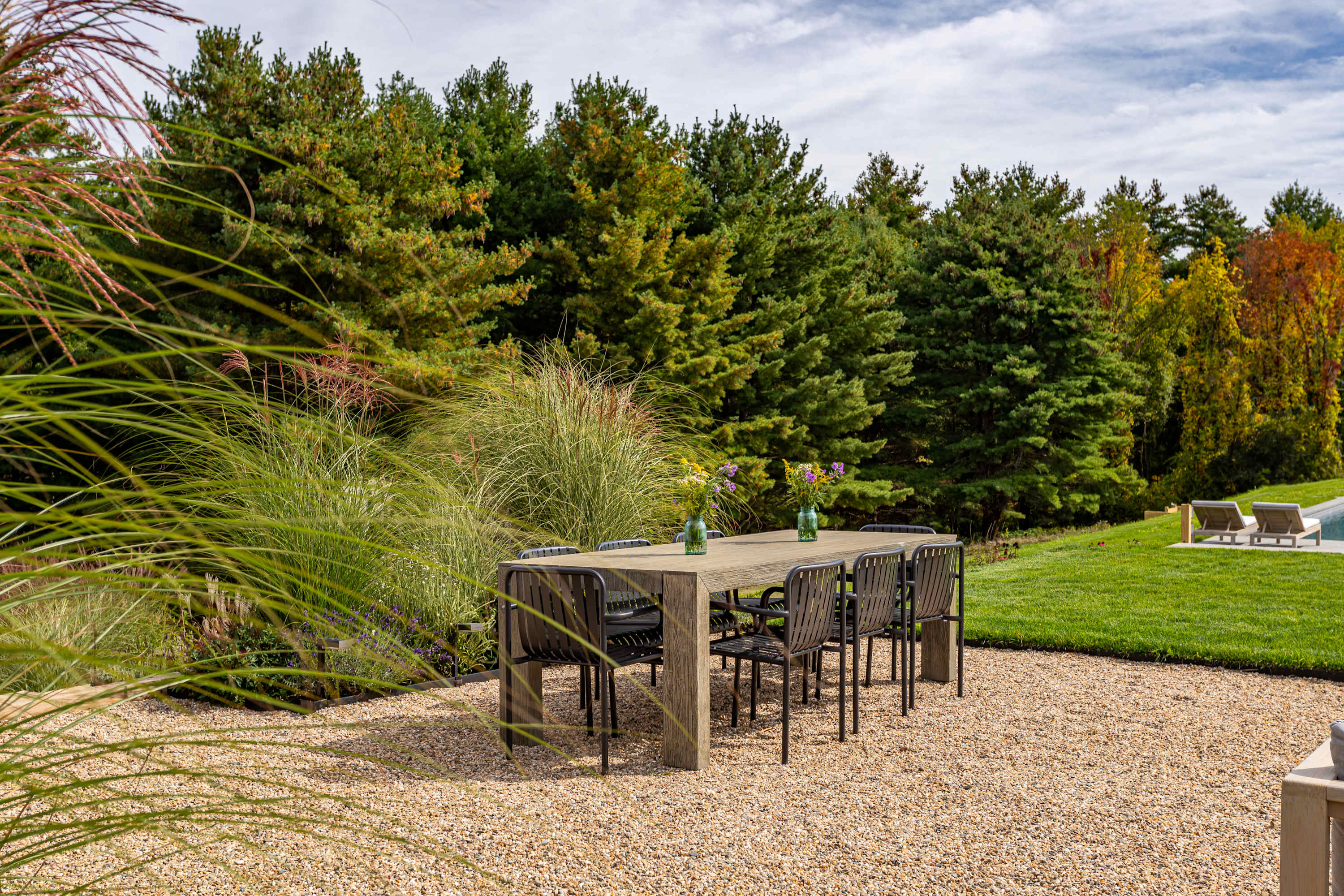 Outdoor dining area featuring a long wooden table with black chairs on a gravel patio, surrounded by tall ornamental grasses and evergreen trees, offering a private and natural setting.