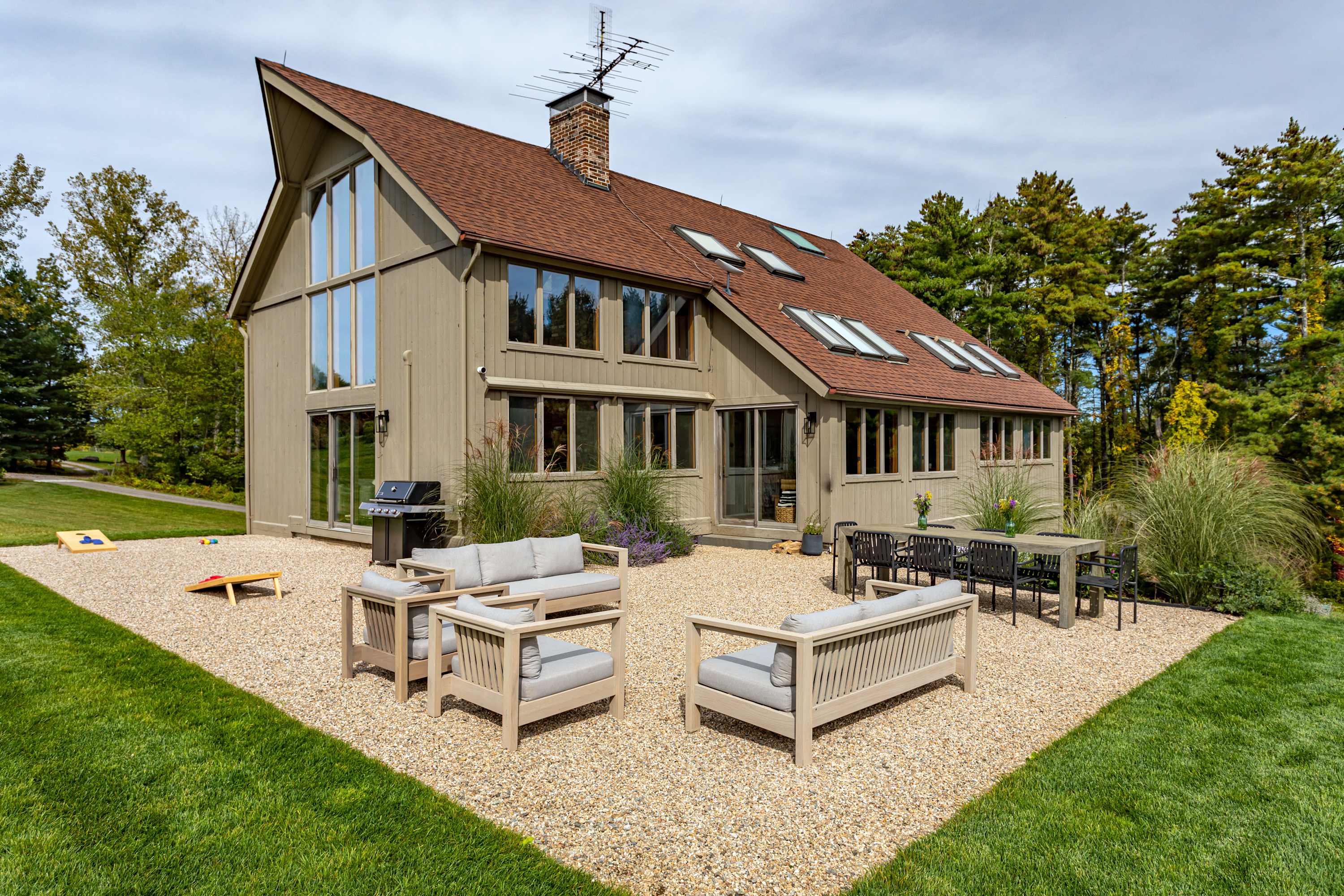 Rear exterior of a modern house with light brown siding and a brown roof, featuring a large gravel patio with outdoor sofas, armchairs, a dining table, and a BBQ grill, surrounded by green lawn and trees.