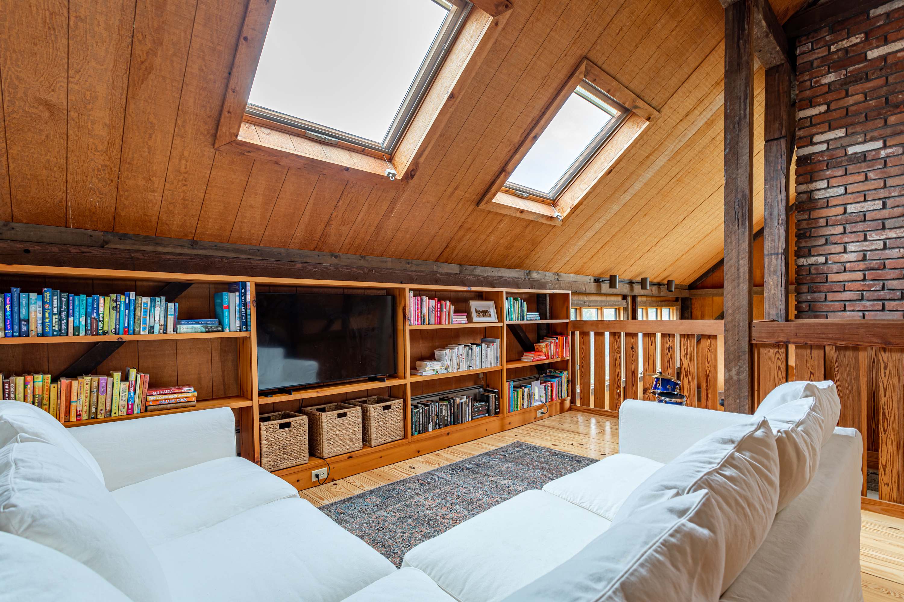Cozy loft living area with two white sofas, a large built-in wooden bookshelf featuring a TV, and a sloped wood-paneled ceiling with two skylights. A patterned rug sits on the wooden floor.