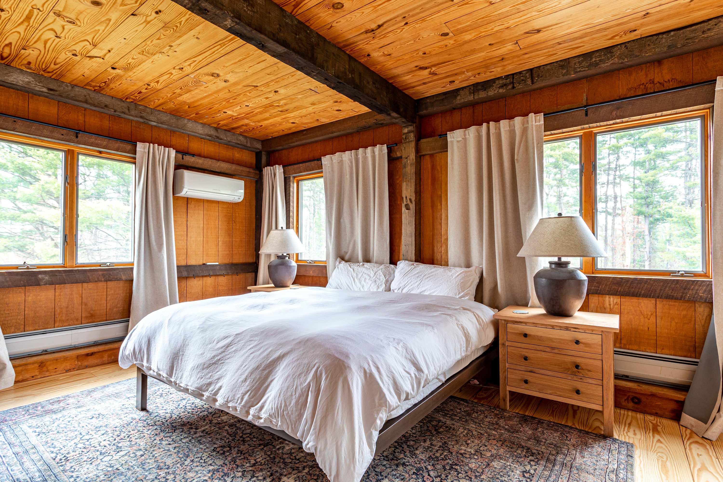 A bright bedroom features a queen-sized bed with white bedding, positioned between two wooden nightstands with lamps. The room is characterized by its warm wood-paneled walls and ceiling with exposed beams, and multiple windows are adorned with light-colored curtains, offering views of the surrounding trees. An air conditioning unit is mounted on the wall.
