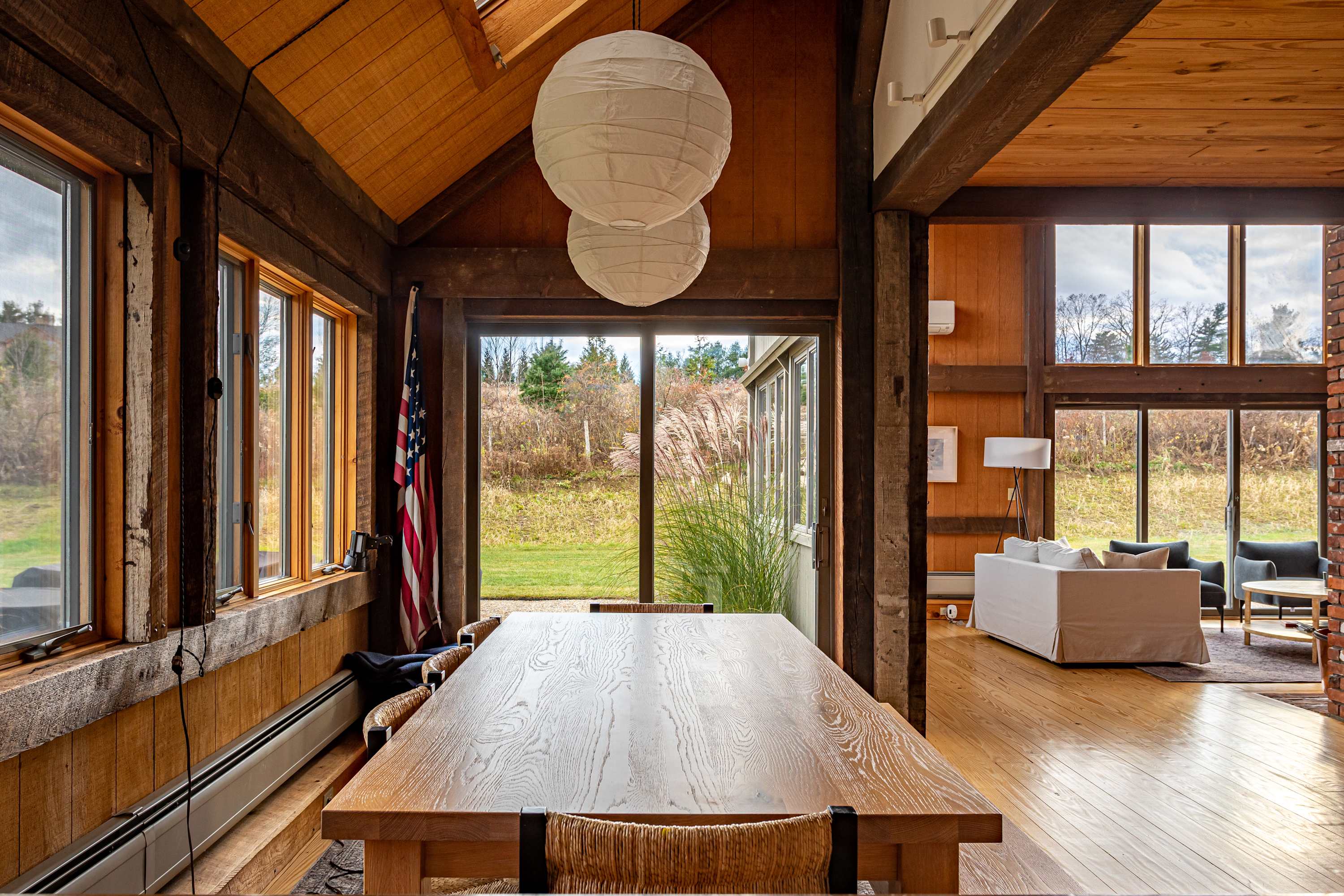A rustic dining area with a long wooden table and chairs, set on a patterned rug, with a view towards the living room through an open archway. Large windows and a sliding glass door provide ample natural light and access to the outdoors. The room features warm wood-paneled walls and a vaulted ceiling with exposed beams, and two large paper lanterns hang above the table.