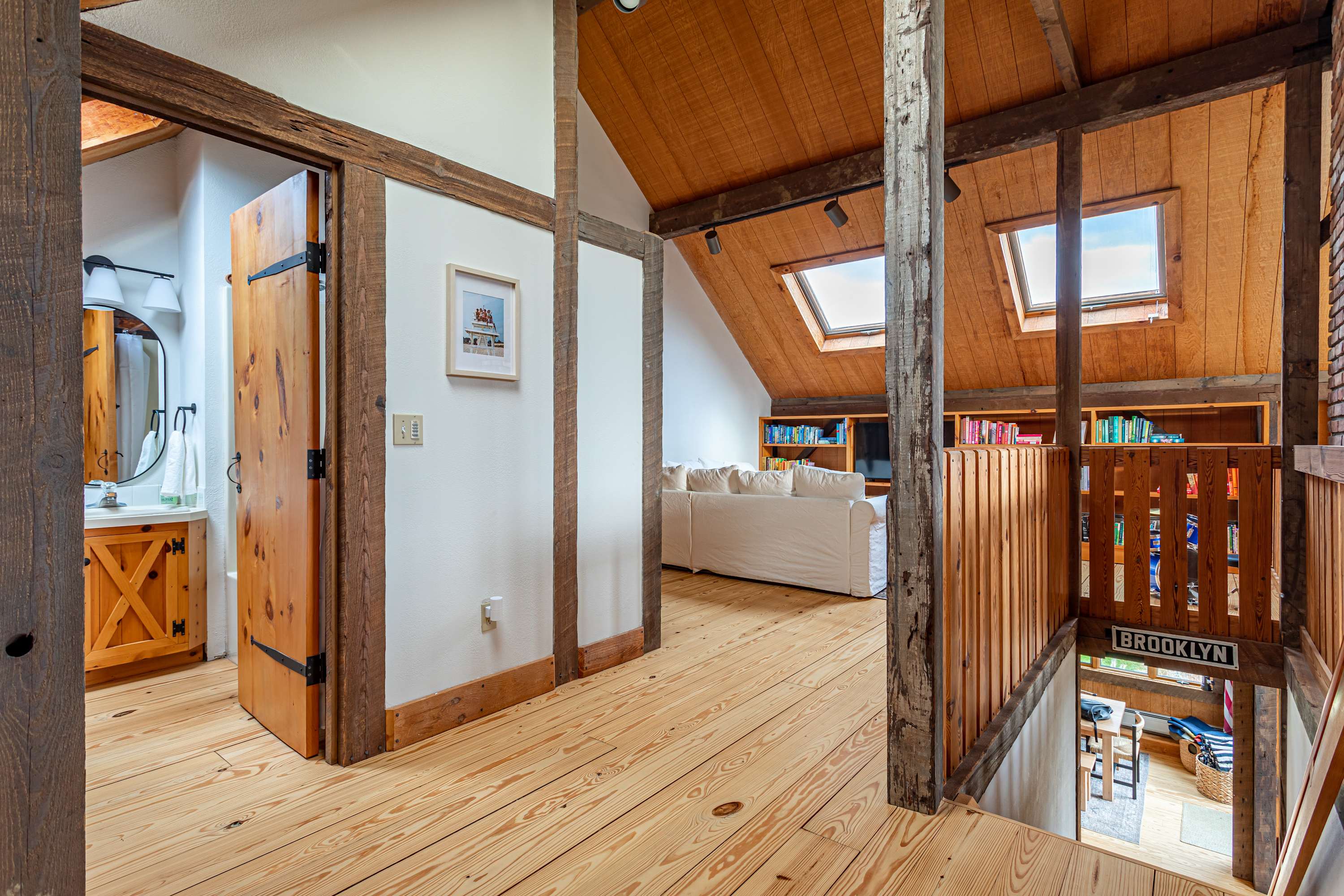 Loft living area with a white sofa, built-in bookshelves filled with books and a TV, and two skylights. Wooden floors and exposed beams add to the rustic charm.
