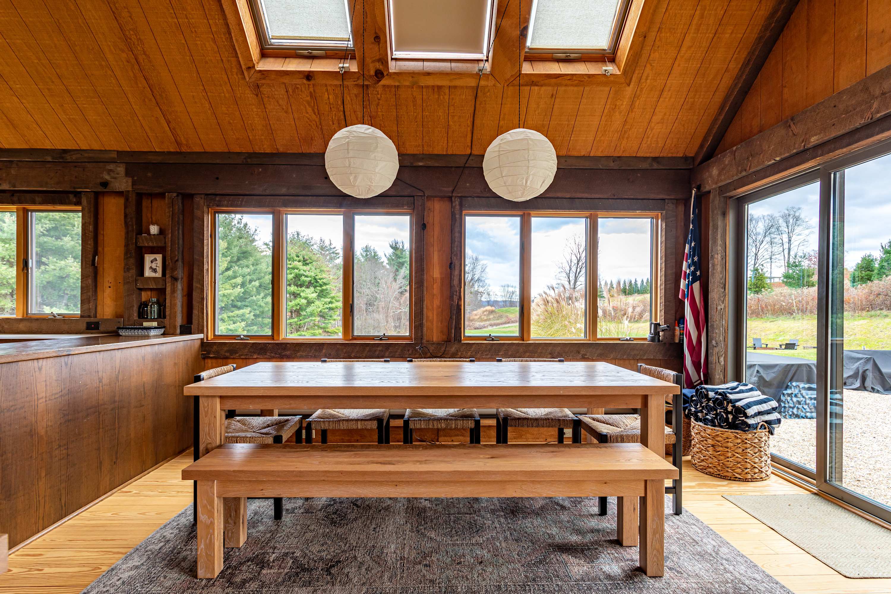 A bright dining area features a long wooden table with benches and chairs, positioned on a patterned rug beneath two large paper lanterns. Expansive windows overlook a lush outdoor landscape, while the room is characterized by its warm wood-paneled walls and vaulted ceiling with skylights. An American flag is displayed near the sliding glass door.