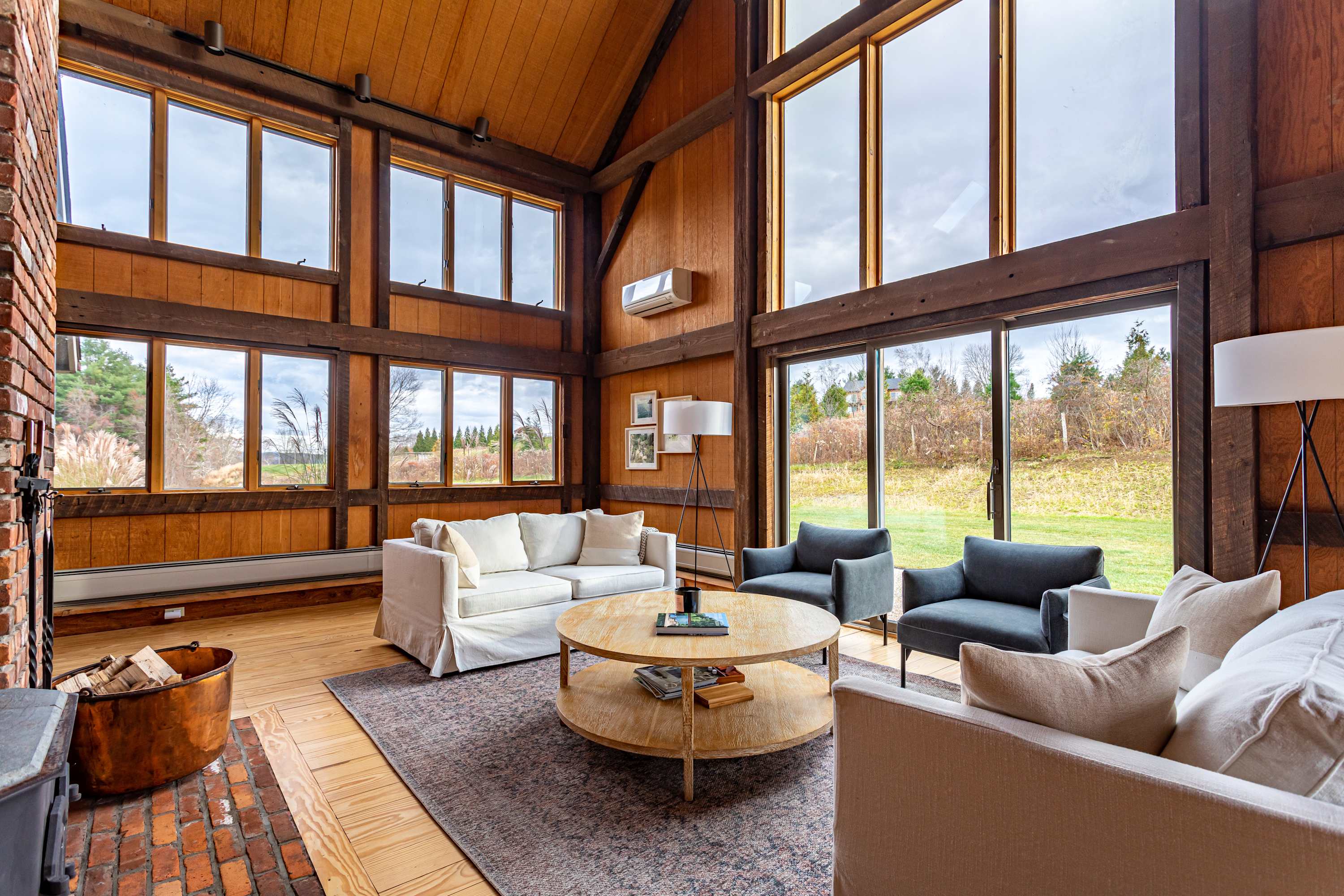 Expansive living room with soaring vaulted ceilings, exposed wooden beams, and a prominent brick fireplace, furnished with comfortable white sofas and gray armchairs on a patterned rug, with large windows providing natural light.