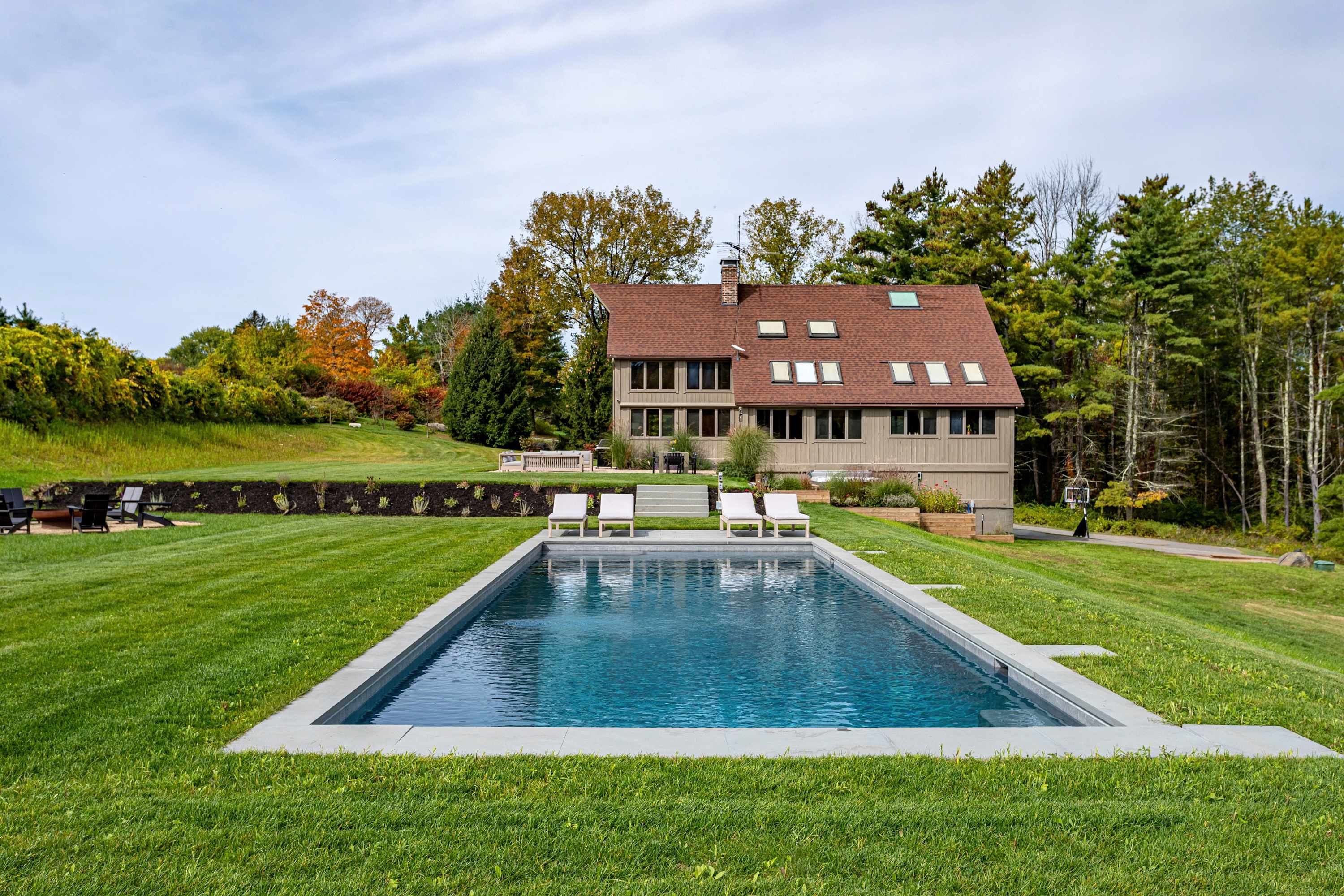 View of the rectangular swimming pool with lounge chairs on a green lawn, leading up to the multi-story house with a brown roof and numerous windows, set against a backdrop of trees and a sloped landscape.