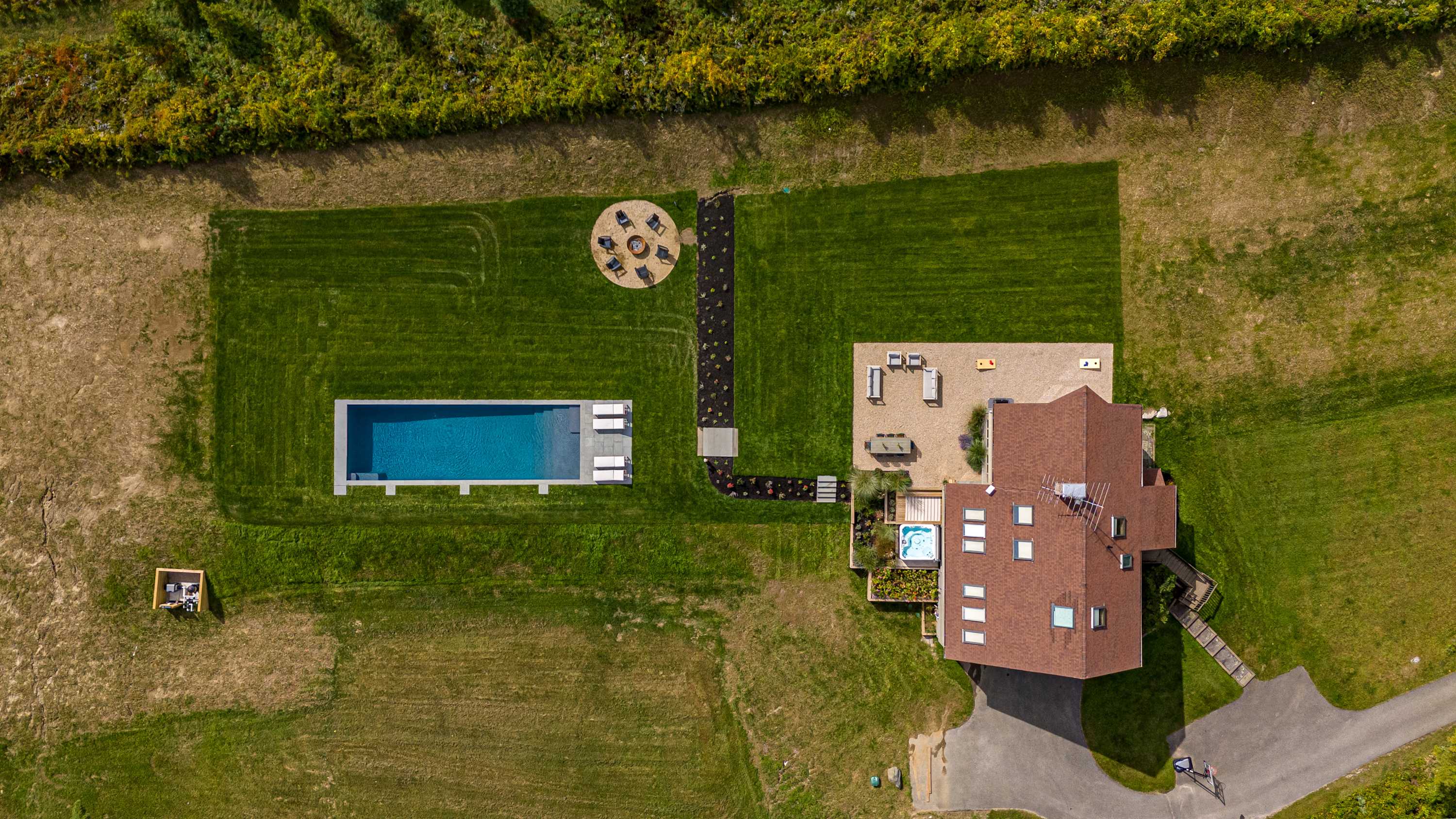 Aerial view of a sprawling property featuring a house with a reddish-brown roof, a rectangular swimming pool with a light-colored deck, a hot tub on a wooden patio, and a circular fire pit area with seating. The property includes extensive green lawns, landscaped paths, and is bordered by dense trees.